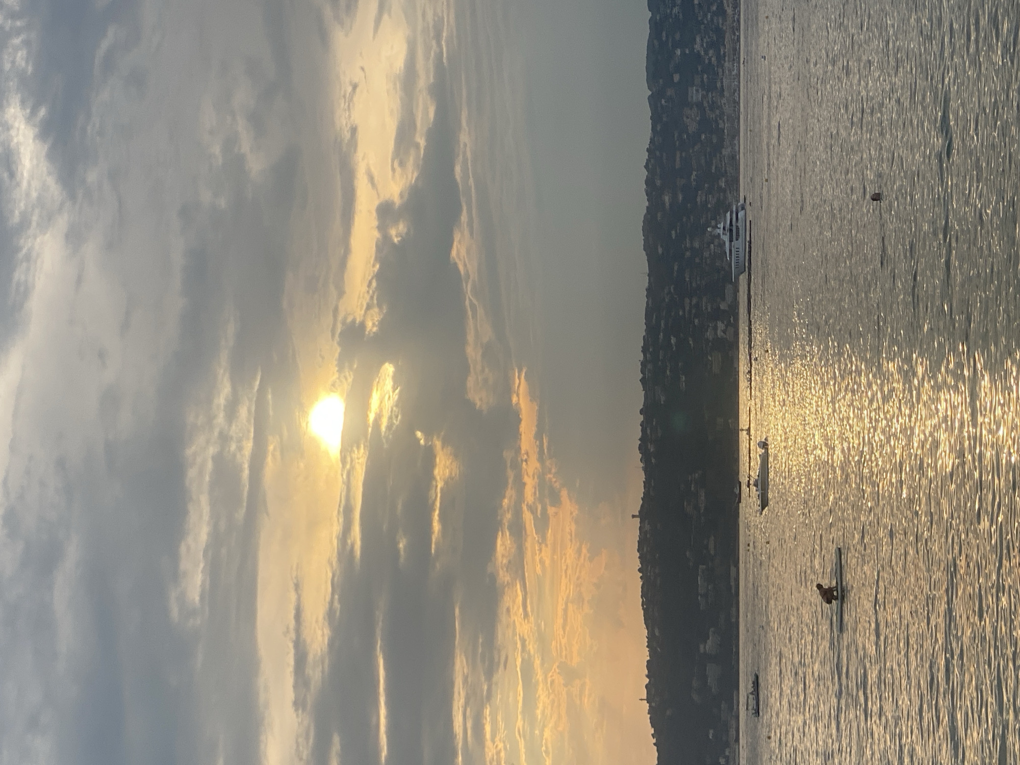 A wide shot captures a sunset over the sea in Antibes, France, with a person on a paddleboard in the foreground. The sky is filled with dramatic clouds, with the sun breaking through and casting a golden glow on the water. A lush, green hillside dotted with houses and buildings forms the backdrop, and several boats, including a large yacht, are visible on the water. The overall scene depicts a serene evening at sea.
