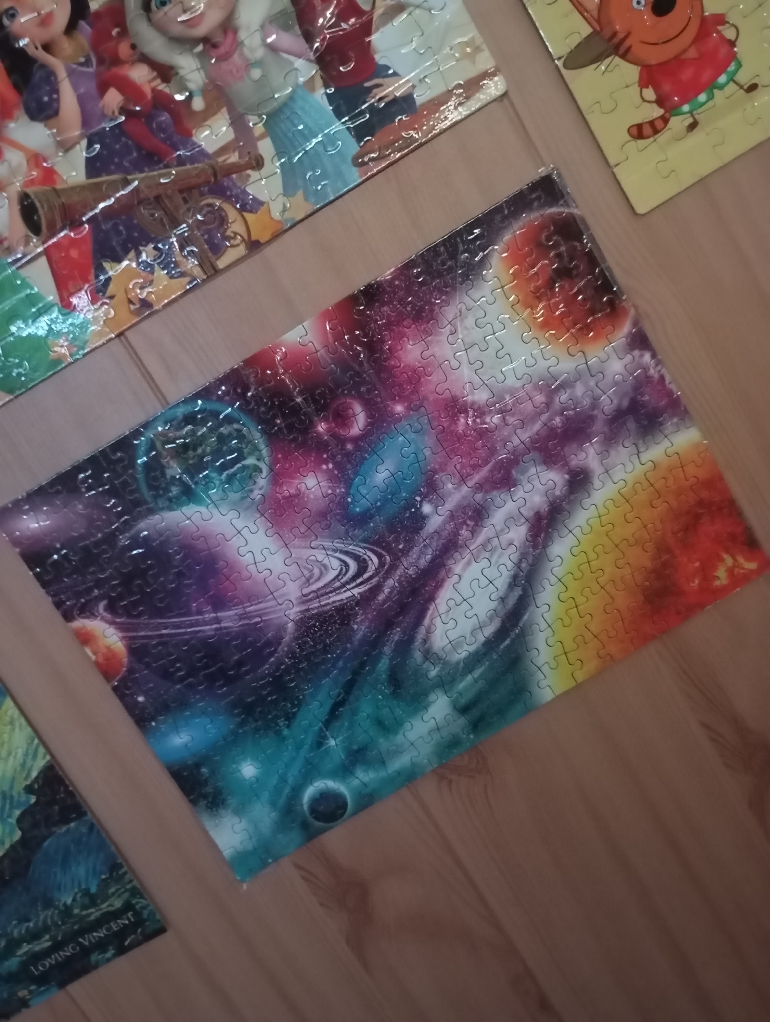 The image shows several completed jigsaw puzzles on a wooden surface. The puzzles depict diverse subjects: cartoon characters, a celestial space scene with planets and nebulae, and a painting with the words "Loving Vincent" visible. The photograph is taken from a slightly elevated angle, capturing the arrangement of the puzzles on the wooden floor.