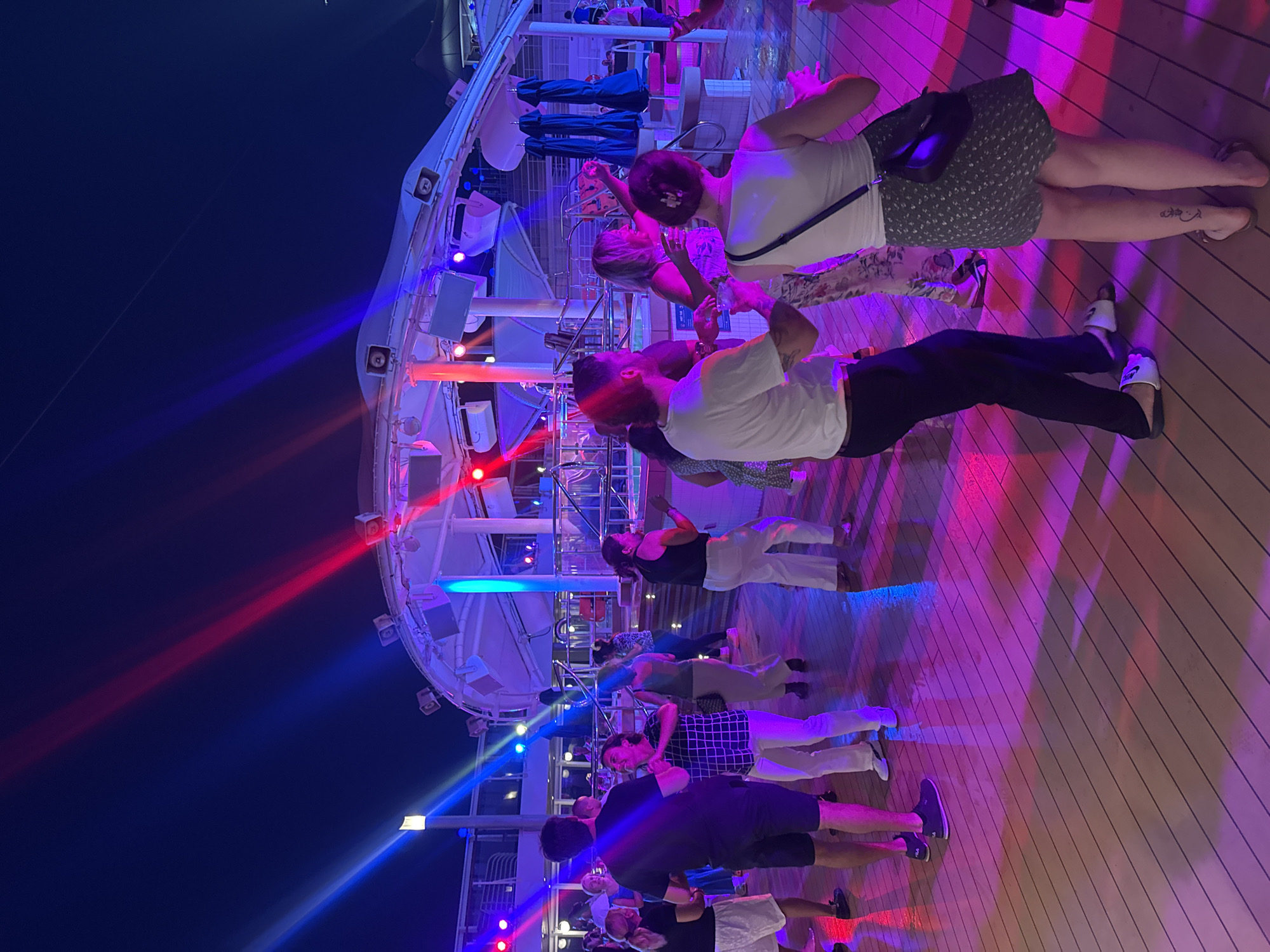 This is a photograph of a nighttime outdoor party or dance event on a wooden deck, likely aboard a cruise ship or at a resort, given the presence of deck chairs and umbrellas in the background. A group of people are dancing and socializing under colored lights, with beams of blue and red light illuminating the scene. The overall atmosphere is festive. The image does not provide any information to suggest a location in Salobreña, Spain.
