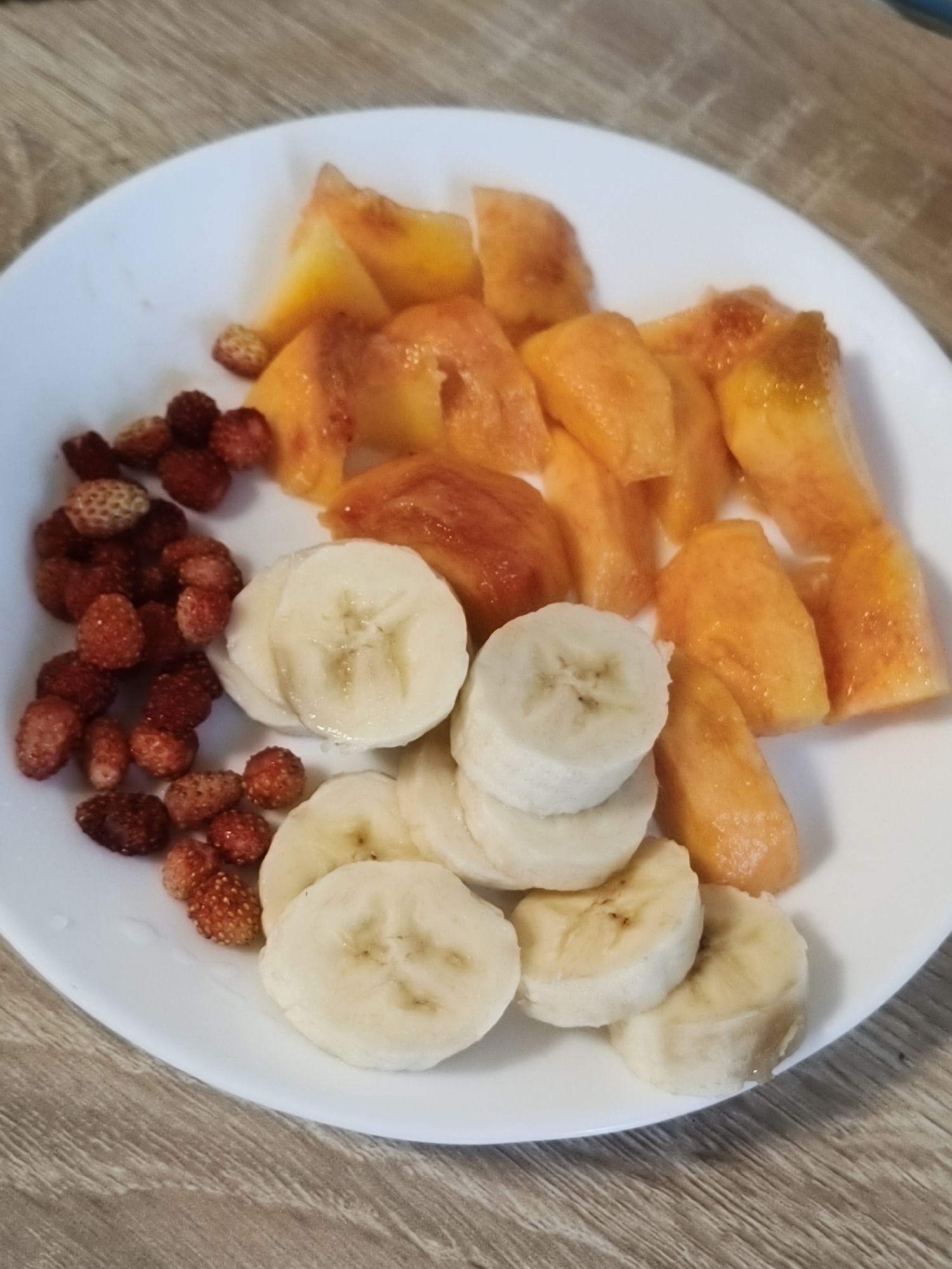 A white plate sits on a wooden surface. The plate is filled with sliced fruit. On the left side of the plate, there is a pile of small, red wild strawberries. To the right of the strawberries are several slices of banana, stacked in two rows. The banana slices are round and white with small brown seeds. The rest of the plate is filled with sliced peaches. The peaches are orange and yellow and appear to be slightly softened. The background is blurred, but it appears to be a wooden table. The image is well-lit and the colors are vibrant.