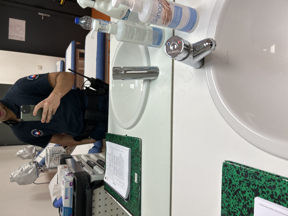 A man in a uniform holds a smartphone, presumably taking a selfie, in front of a white sink and counter. On the counter are soap bottles, a small water bottle, and a chrome faucet. A clipboard with papers is attached to the cabinet below the sink. Next to the counter is a cart with medical supplies. A small portion of a green folder with black spots is visible on the right. The image is taken at an unusual angle.