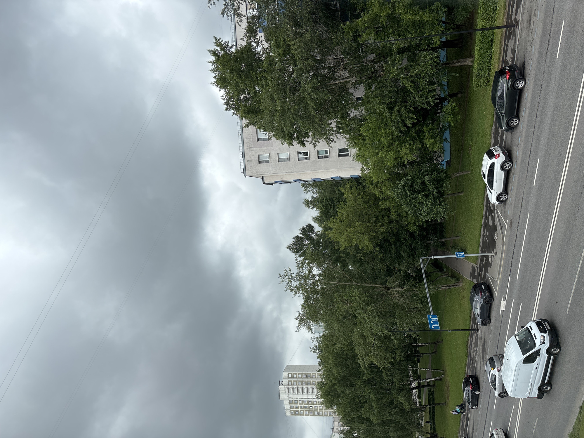 The image captures a street scene in Khimki, Russia, on a cloudy day. A multi-lane asphalt road runs diagonally across the frame, with several vehicles present. A white van is in the foreground, heading away from the viewer. Behind it, a silver hatchback is visible. Further up the road, a dark SUV is parked, and a white SUV is in the lane closest to the sidewalk. On the sidewalk, two figures are walking with a stroller. Adjacent to the road is a green grassy area with trees and a sidewalk. In the background, two multi-story buildings are visible, one taller and white with many windows, and another lighter-colored building partially obscured by trees. Power lines are strung across the overcast sky. The overall impression is an urban environment with elements of nature.