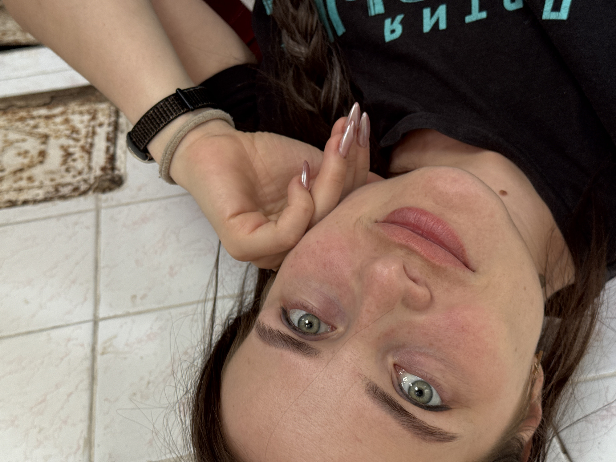 A woman is lying down, looking up at the camera. She has fair skin, green eyes, and light pink lips. Her hair is dark and partly covers her shoulder. She wears a black t-shirt with blue lettering. Her left hand is raised near her face, revealing long, iridescent nails. On her left wrist, she wears a black smart watch and a light-colored elastic bracelet. The background consists of a tiled floor and a patterned rug.
There is no information to determine the location in the city of Rusalevka, Russia.