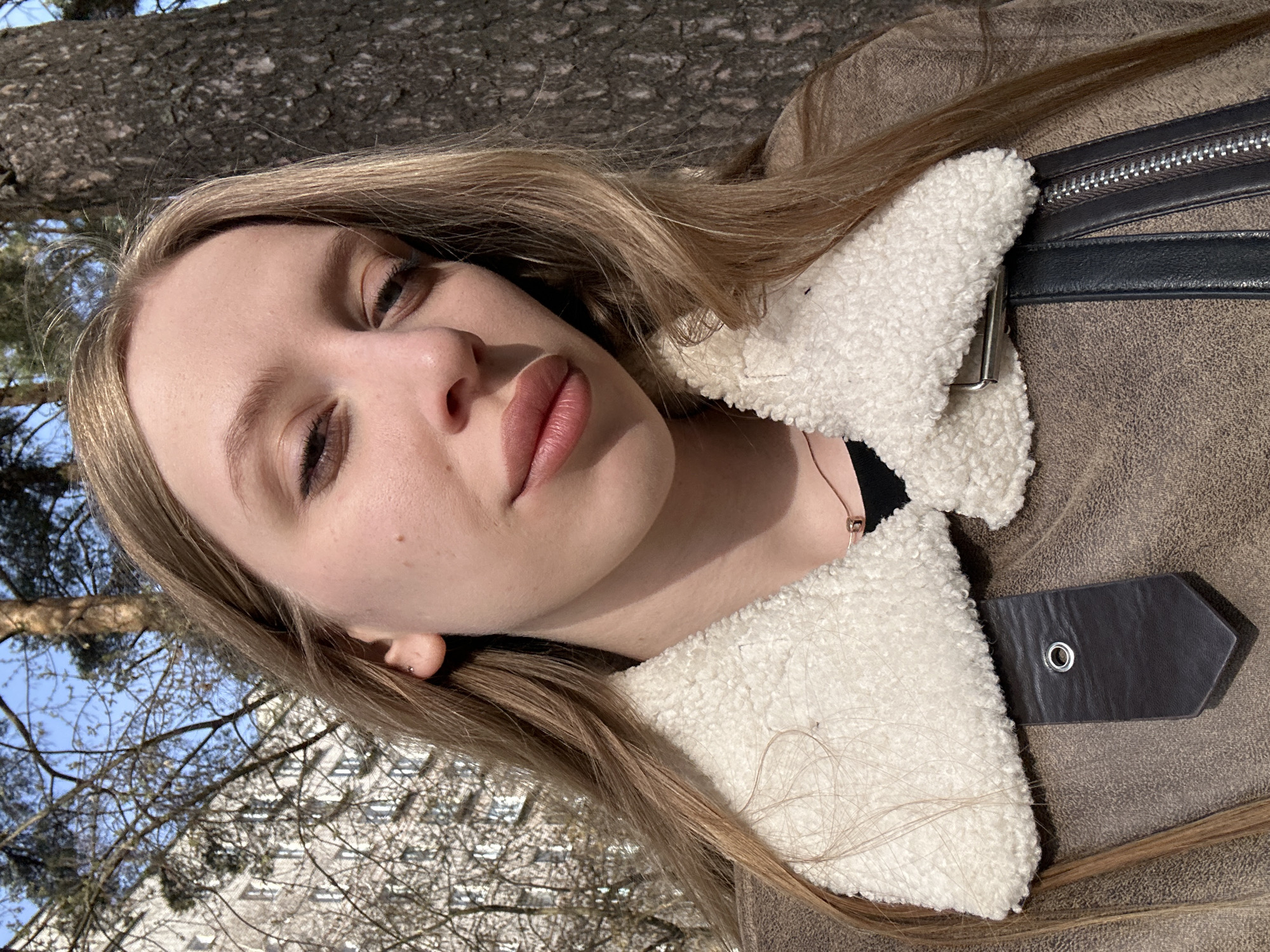 A young woman is seen in a tilted close-up shot. She has fair skin, light brown hair, and is wearing a brown jacket with a white, fluffy collar. The background reveals a tree and a building, suggesting an outdoor setting possibly in the city of Chaikovsky, Russia.