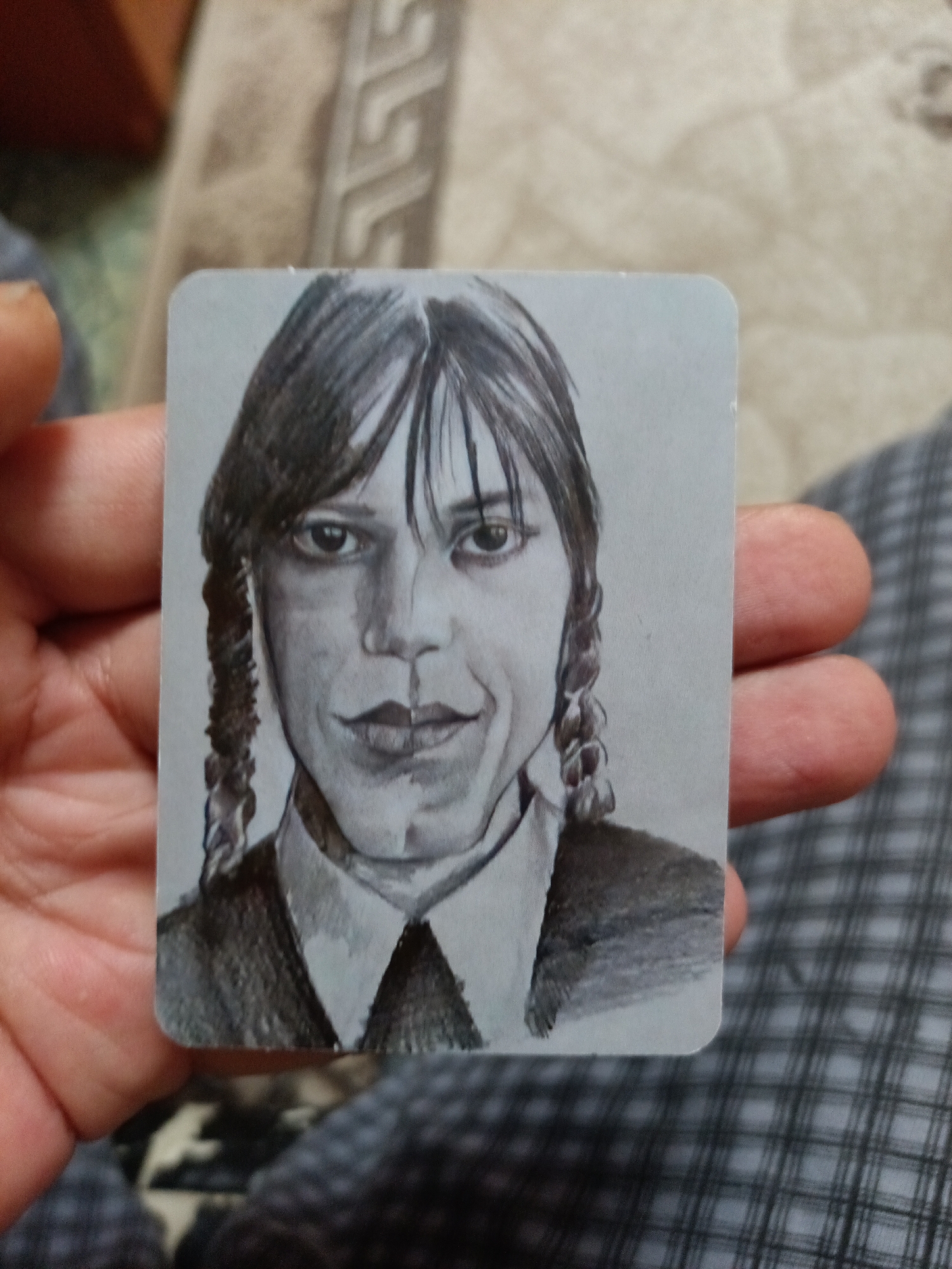 The image shows a grayscale drawing of Wednesday Addams, rendered in pencil. The drawing is on a rectangular card with rounded corners, and it is held by a person's hand. The person's thumb and index finger are visible on the left side of the card, and their other fingers are on the right side. The background is blurry and appears to be a patterned fabric, possibly a blanket or upholstery. There is no indication of the city of Nyagan, Russia, or any urban environment in the image. The drawing itself depicts Wednesday Addams with her signature braided pigtails, bangs, and a stern expression. She is wearing a collared shirt and a dark top. The drawing is detailed, with shading used to create depth and form.