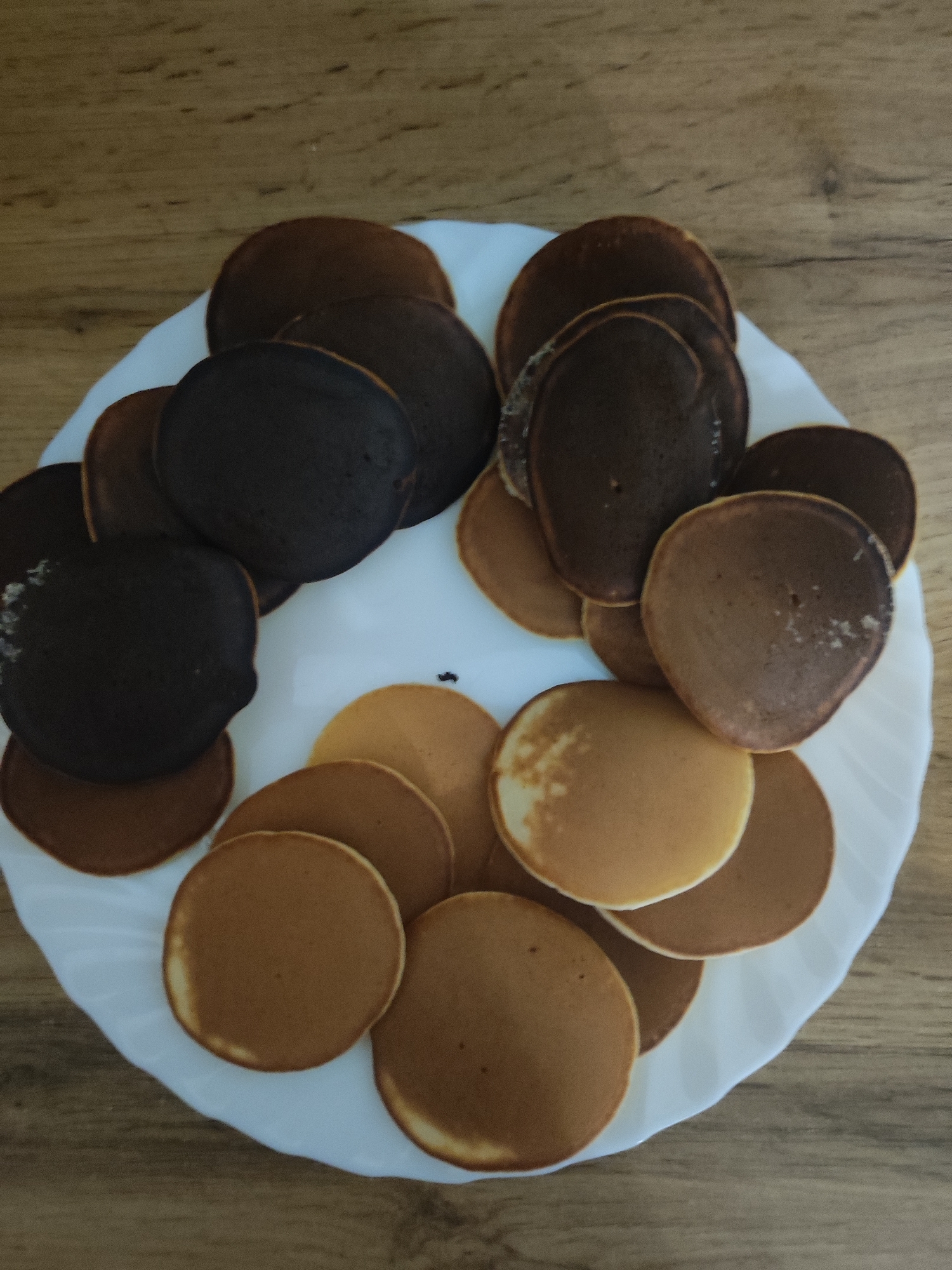 On a wooden surface, a white plate is piled with a collection of circular pancakes. The pancakes vary in color, ranging from light brown to dark brown, some appearing burnt. There are no people visible in the image, but it is likely that they were the ones who prepared the pancakes. The location is not evident from the image.