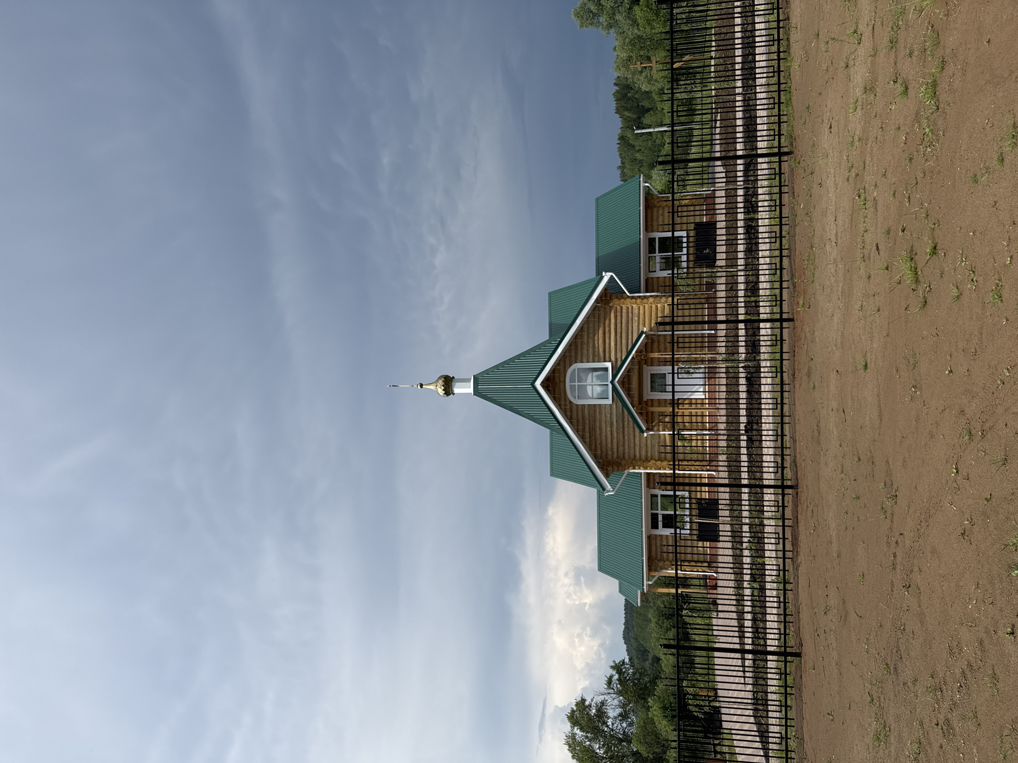 A wooden Orthodox church with a green roof and a golden dome stands behind a black metal fence. The church is made of logs, with white trim around the windows and doors. The fence encloses a courtyard with a brick walkway and some landscaping. In the background, there are trees and hills, and the sky is cloudy. The scene appears to be in Вассята, Russia.