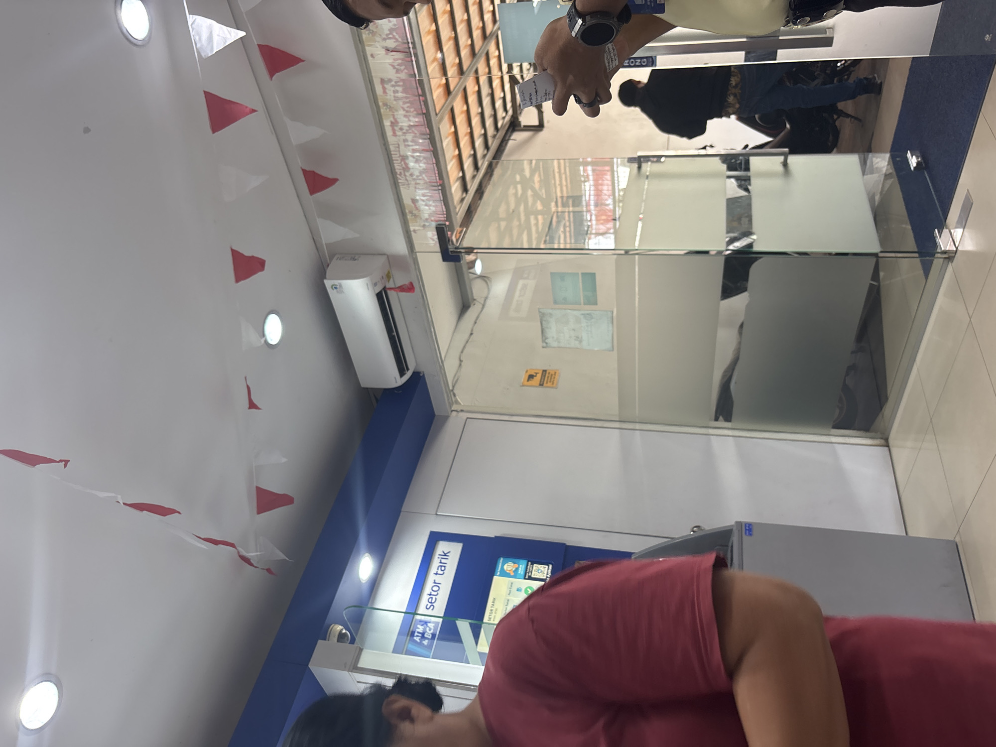 The image shows the interior of a bank in North Jakarta, Indonesia, possibly a BCA bank, given the "ATM BCA" signage visible on an ATM machine. The scene includes an ATM machine with "SETOR TARIK" (Deposit Withdrawal) written on it. Red and white triangular flags are strung across the ceiling, suggesting a national celebration or holiday. Several people are present, including a woman in a red shirt in the foreground, a person in a black jacket and jeans reflected in a glass partition, and another person holding a piece of paper and a watch on their wrist. The environment is brightly lit with ceiling lights and an air conditioning unit is visible. The overall impression is a public space, likely a bank branch, decorated for an event.