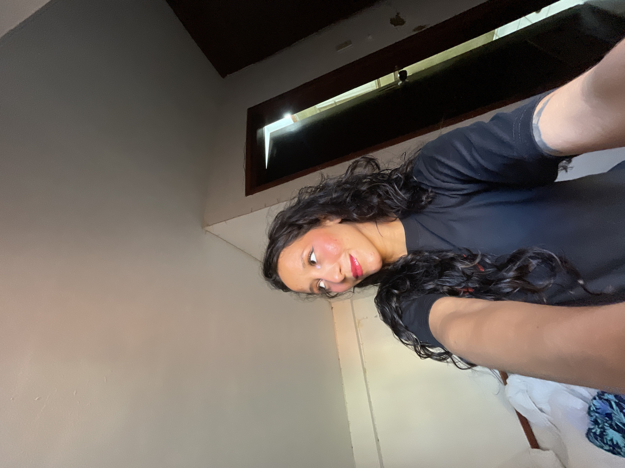 The image shows a woman with dark, curly hair wearing a black t-shirt. She is looking to her right and has bright pink blush on her cheeks and red lipstick. Her right arm is extended forward, holding a camera or phone, as if taking a selfie. The background appears to be an indoor setting, possibly a room, with a partially open dark wooden door revealing light from an adjacent area. The walls are a light color, and there is a bed with white linens visible to the left of the woman. The lighting suggests it is daytime, with light entering from the side and through the open doorway. This scene is located in Porlamar, Venezuela.