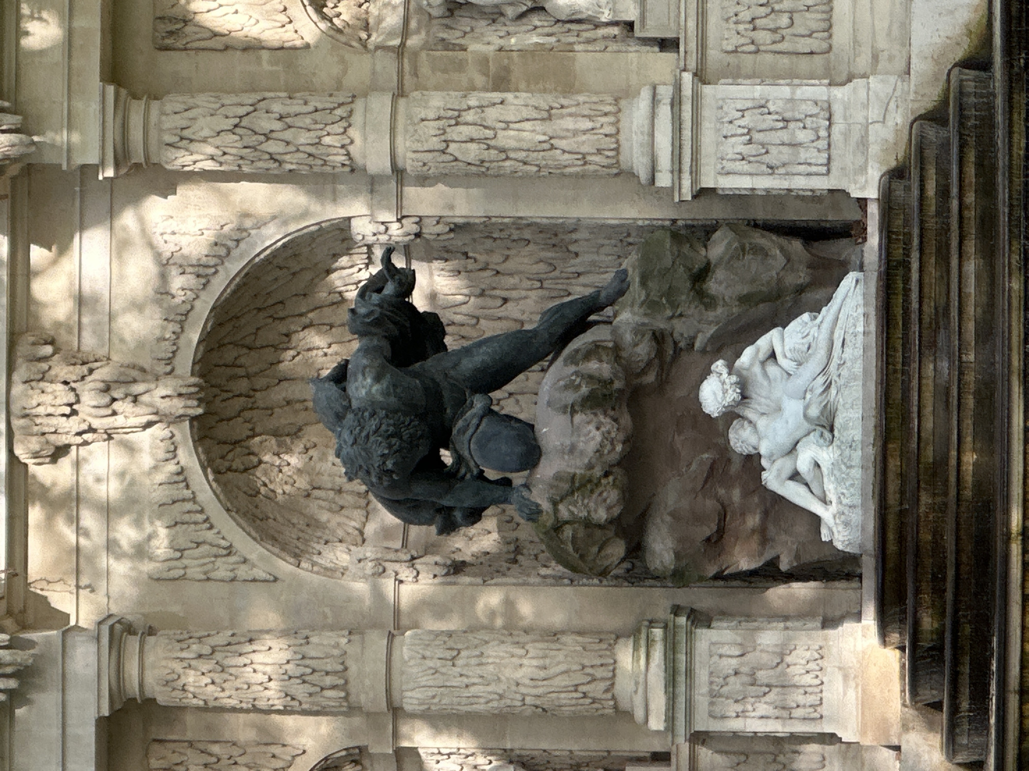 The image depicts the Fountain of the Medici in the Jardin du Luxembourg, Paris, France, seen from an unusual, nearly vertical perspective. The fountain features a dark bronze sculpture depicting a male figure (possibly a satyr or faun) wrestling or grappling with a lion or similar animal, mounted on rough rocks. Adjacent to this sculpture is a light-colored marble sculpture of a reclining figure, possibly a nymph, and a smaller human figure. The sculptures are set within a shallow arched alcove. The background consists of a textured stone wall with pilasters and molding. To the right are steps constructed with vertically aligned wooden beams. The scene is bathed in dappled sunlight, casting shadows across the stone surfaces.