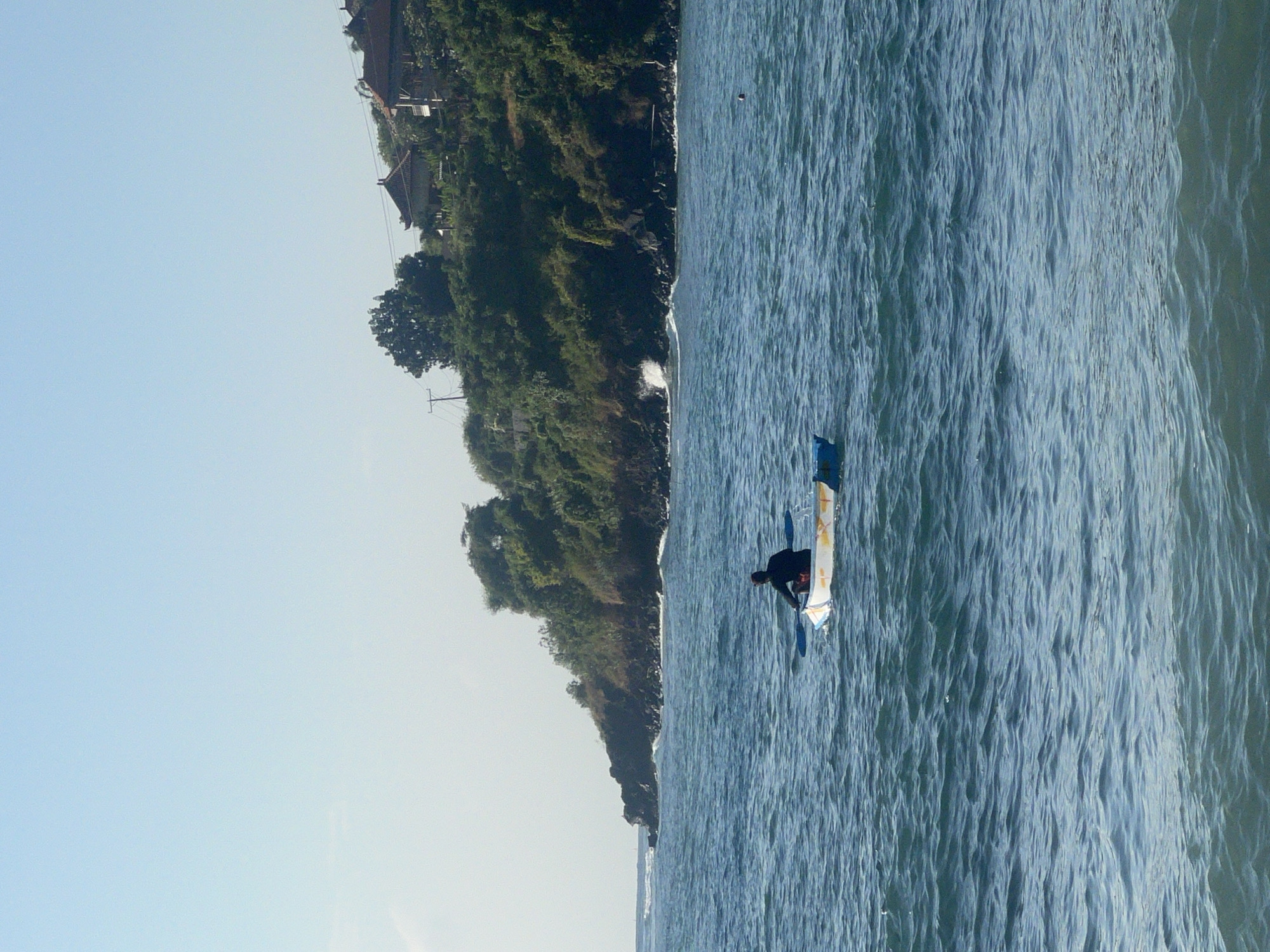 In the city of Abang, Indonesia, a single individual is seen kayaking on the bright blue ocean under a clear sky. The kayaker, wearing a dark shirt, is paddling a small, white and yellow kayak. The shoreline in the background is lined with lush green trees and several buildings with red roofs, suggesting a residential or resort area. Waves gently break against the rocky shore. The scene is tranquil and highlights the natural beauty of the coastal landscape.