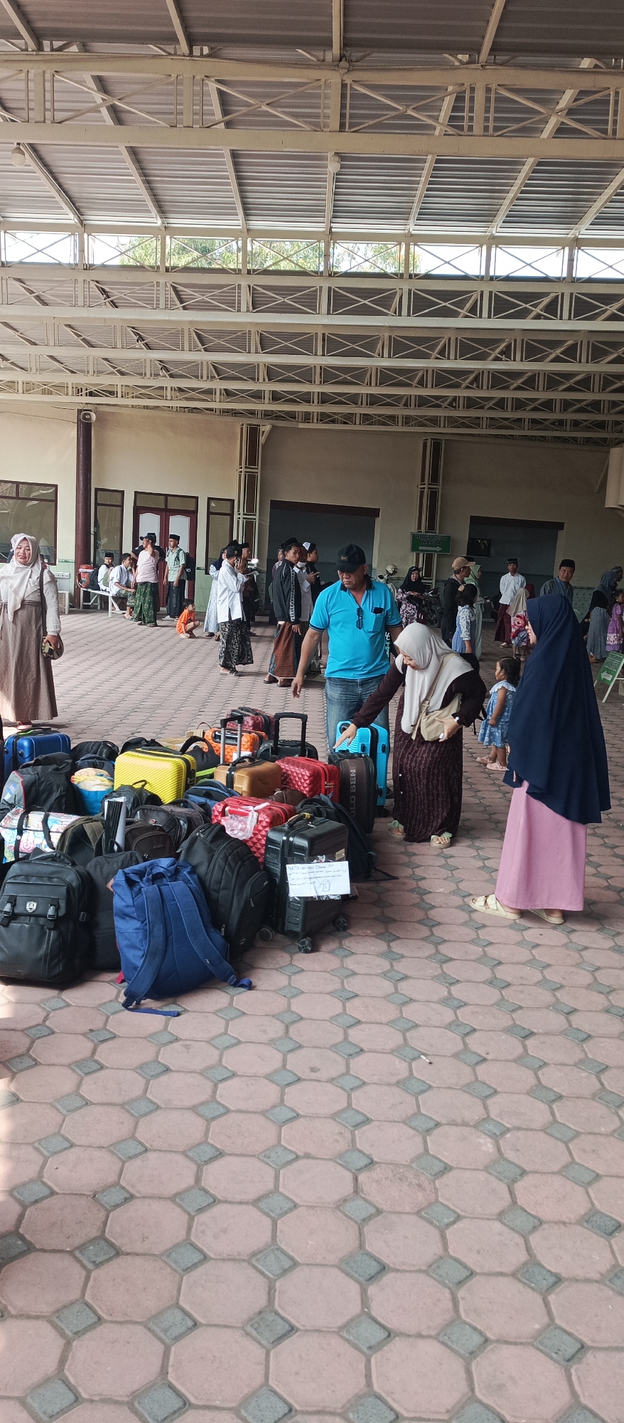 This image depicts a gathering of people and luggage at what appears to be a transportation hub in Kapongan, Indonesia. The scene is set under a large, open-air roof supported by metal beams, suggesting an outdoor waiting area or terminal. The ground is paved with octagonal and diamond-shaped tiles.
In the foreground, a large collection of various luggage items, including suitcases, backpacks, and duffel bags, are piled together. Several people are interacting with or standing near the luggage. A man in a blue t-shirt and jeans is looking towards the luggage, with his hand pointing. To his right, a woman in a dark hijab and a pink skirt stands. To the left of the man, a woman in a light-colored hijab and a long brown skirt is visible.
In the background, a larger group of people are standing or sitting, some appearing to be waiting. Many are dressed in traditional clothing, including headscarves and sarongs. Buildings with windows and doors are visible in the far background. The overall impression is one of travel or departure.