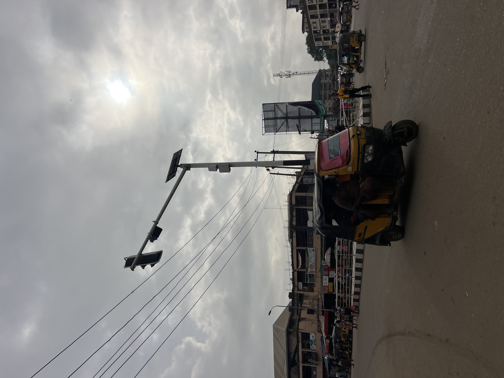 In Nkpor, Nigeria, on an overcast day, a busy street scene unfolds. In the foreground, a yellow and black auto-rickshaw, also known as an "okada" or "keke napep", with a red roof is prominently positioned, carrying at least two passengers. The vehicle is on a paved road, with tire marks visible on the asphalt.
Beyond the auto-rickshaw, a traffic light with three signal heads hangs suspended over the intersection by a metal pole equipped with a solar panel. Several power lines are strung across the sky. In the middle ground, a variety of other auto-rickshaws, some yellow and some blue, are visible, indicating ongoing transportation activity. Pedestrians are also present, some walking, others seemingly waiting.
The background features multi-story buildings lining the street, exhibiting various architectural styles and signs, suggesting commercial establishments. A prominent billboard stands tall, and a cell phone tower can be seen in the distance. The sky is heavily clouded, with the sun partially visible through the clouds, casting a diffused light on the scene. The overall impression is one of a bustling urban environment with active daily life.