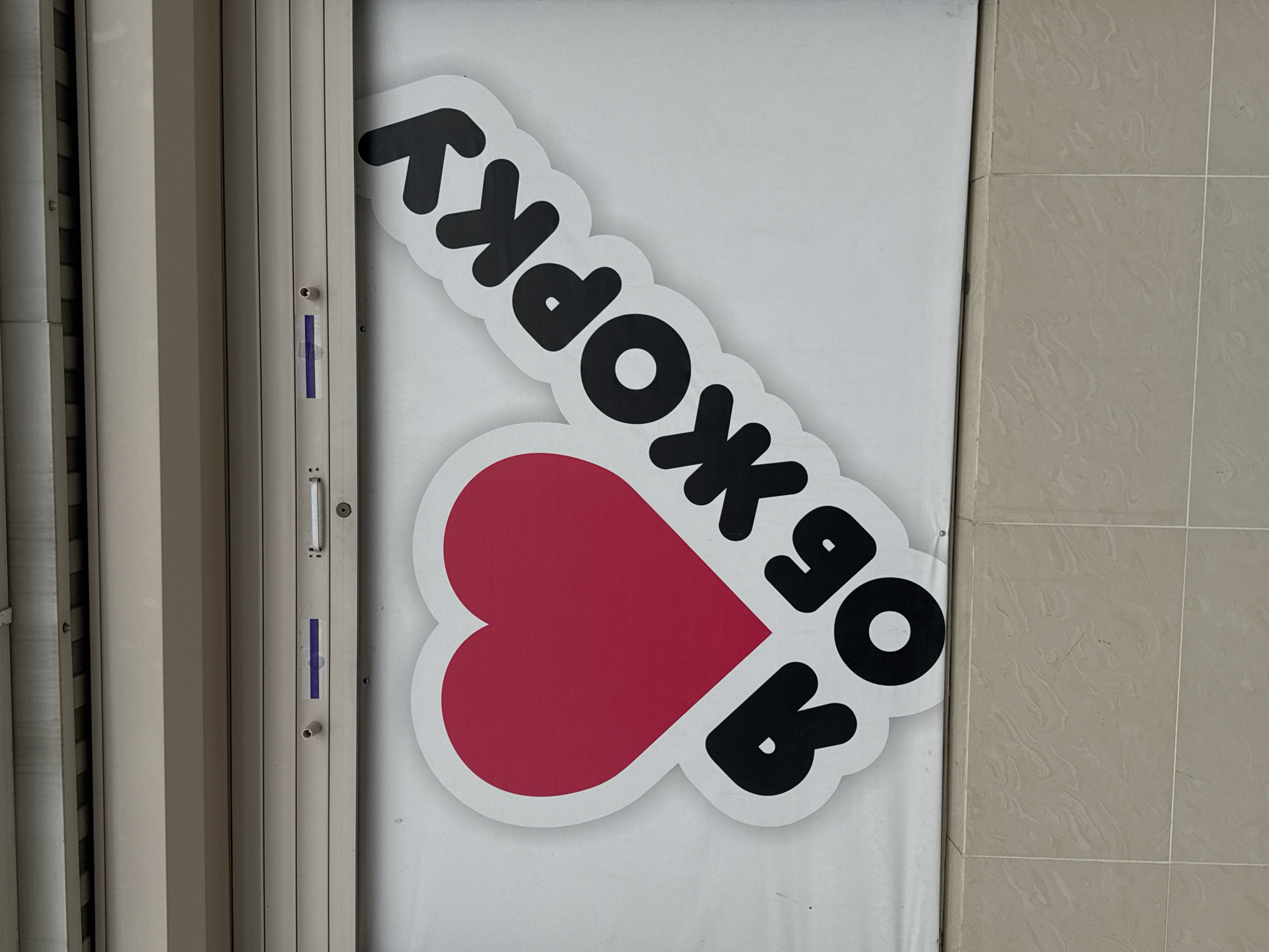 The image shows a sign in Russian on a door, featuring the text "В Художку" (V Khudozhku) above a heart shape, indicating a possible art-related location. The scene is near a tiled wall and a closed shutter door. It could be a store, studio, or school related to art. There is no evidence in the image that identifies the location in the city of Ivanovskaya, Russia.