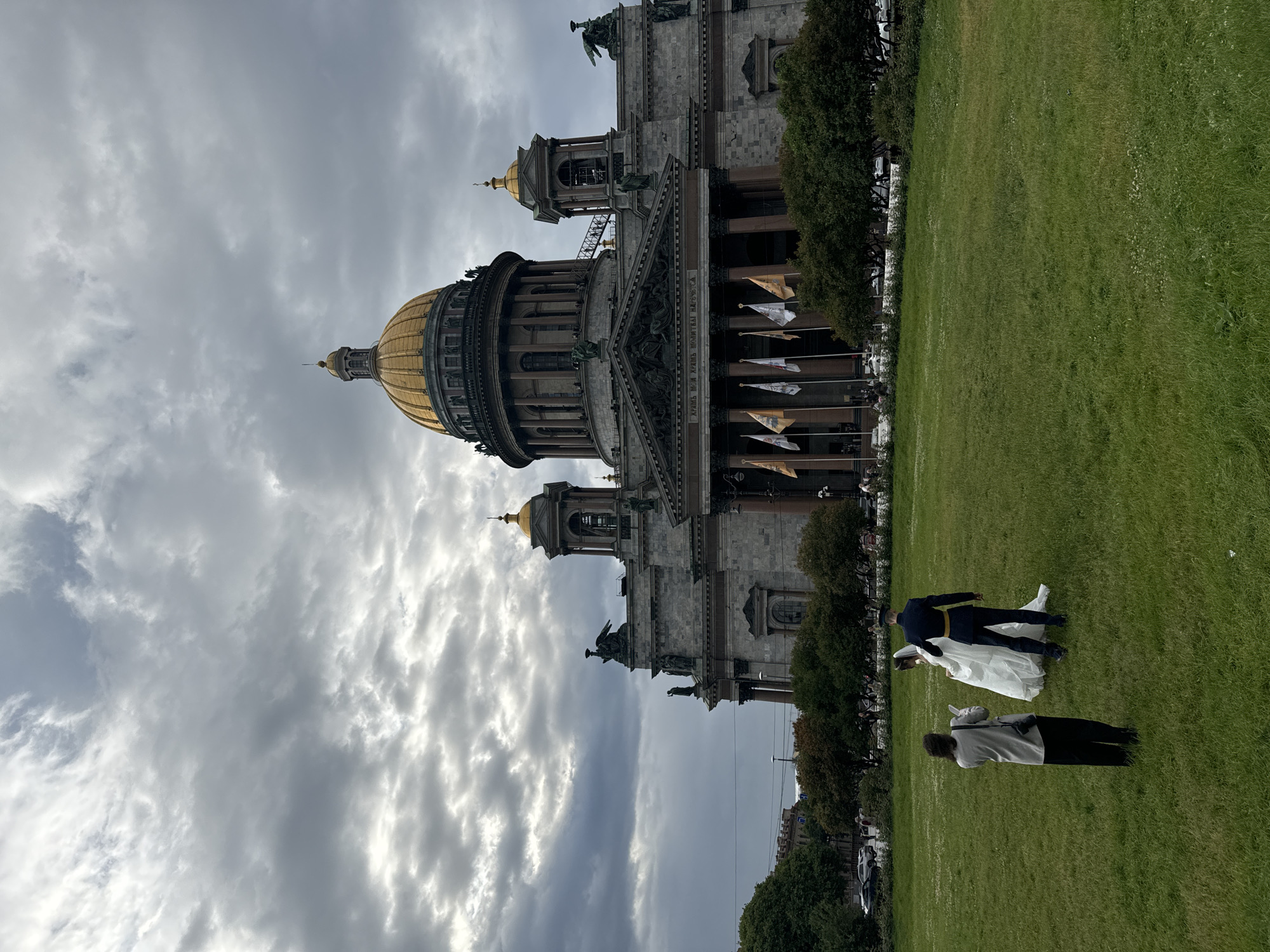 In Saint Petersburg, Russia, under a dramatic, overcast sky, a wedding couple is being photographed on a vast, green lawn in front of Saint Isaac's Cathedral. The bride, in a flowing white gown, stands beside the groom, who is dressed in a dark military-style uniform with a gold sash. A photographer, holding a camera and a smartphone, stands a short distance away, capturing the moment. The magnificent cathedral, with its iconic golden dome, forms a grand backdrop to the scene. Trees line the edge of the lawn, and a street with parked cars is visible in the distance. Flags fly in front of the cathedral, adding to the solemnity of the occasion.