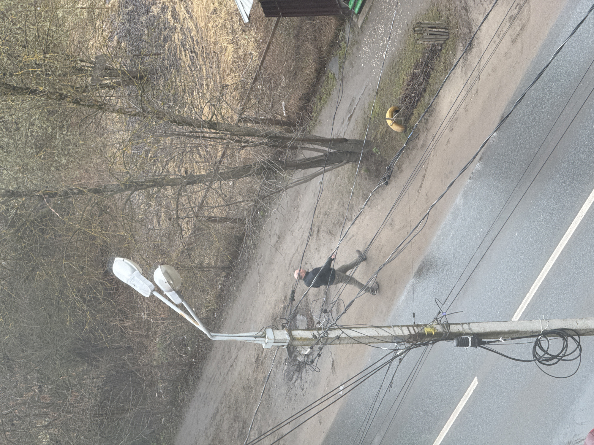 Viewed from above, the scene shows an outdoor location. A man in dark clothing and light-colored pants lies on a dirt path near an electrical pole. The man appears to be lying between the pole and a tree, and the power lines run across the path. A street runs parallel to the path. A tire hangs on a pole next to the path. The location is inferred to be Tver, Russia, based on the prompt.