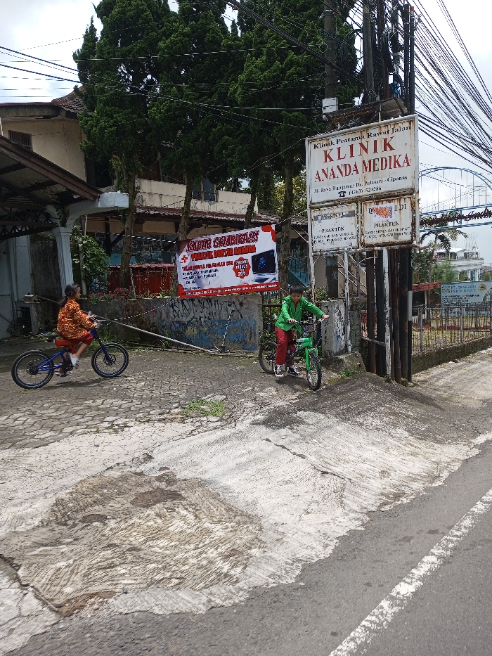 This image captures a daytime street scene in the city of Chendenbji, Bhutan. Two individuals, identified as children, are present on bicycles on a paved surface. The child on the left, wearing an orange and brown patterned shirt and dark trousers, rides a blue bicycle with a red seat, facing right. The child on the right, clad in a green shirt and red trousers, is on a green bicycle, positioned facing slightly left near a concrete ramp.
In the background, a light-colored building with a dark roof is partially obscured by several tall, dark green coniferous trees. To the right, a prominent white sign, mounted on a utility pole, displays "KLINIK ANANDA MEDIKA" along with additional Indonesian text detailing clinic services, a doctor's name, and a phone number. Below this sign, a red and white banner with Indonesian text and a computer monitor graphic advertises "JALUR SEMBILAN" and "SERVICE LAPTOP MOTHERBOARD." Numerous overhead utility wires are visible across the upper portion of the frame. The ground transitions from irregular grey paving stones to a patched concrete ramp, leading down to an asphalt road with a white painted line on its right edge.