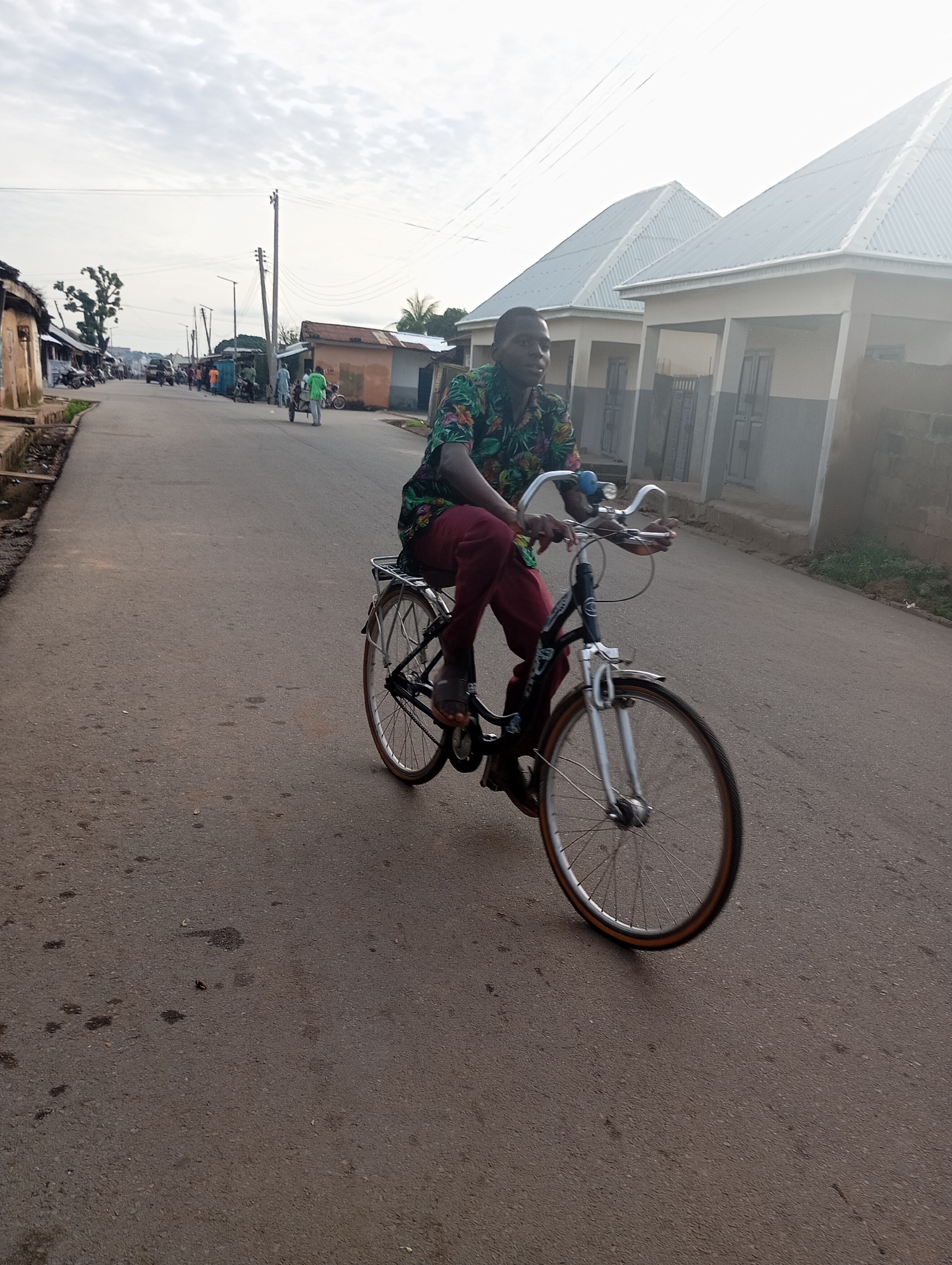 The image depicts a young Black male riding a bicycle on an asphalt road in Shendam, Nigeria. He is wearing a colorful floral print shirt and maroon pants, and is on a blue bicycle with a silver rack on the back. The road is lined with buildings, some appearing to be houses with corrugated metal roofs and others with more traditional architecture. There are other people and vehicles visible in the background, suggesting a populated area. The sky is overcast with a bright horizon. The scene appears to be in a residential or semi-commercial area of the city.
