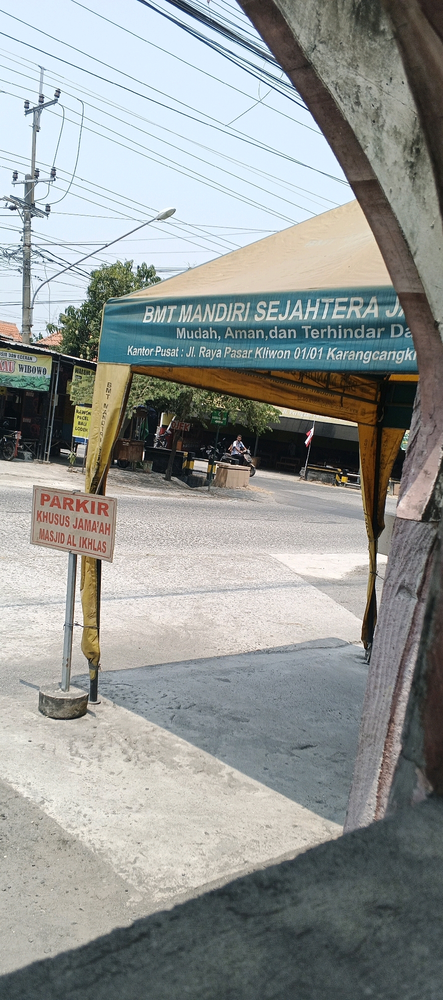 The image is a medium shot of a street scene in Sumberwudi, Indonesia. The foreground is dominated by a concrete sidewalk and a parking sign. The sign is red and white and reads "PARKIR KHUSUS JAMA'AH MASJID AL IKHLAS", indicating parking is reserved for mosque attendees. Behind the sign, a yellow and blue tent is set up, likely for an event or vendor. Text on the tent reads "BMT MANDIRI SEJAHTERA JA" and "Mudah, Aman, dan Terhindar Da" and "Kantor Pusat: Jl. Raya Pasar Kliwon 01/01 Karangcangk". Several people are visible in the background, including a person riding a motorcycle. The street is paved and there are trees and buildings lining the street. Overhead, power lines and a street lamp are visible. The sky is clear and blue.