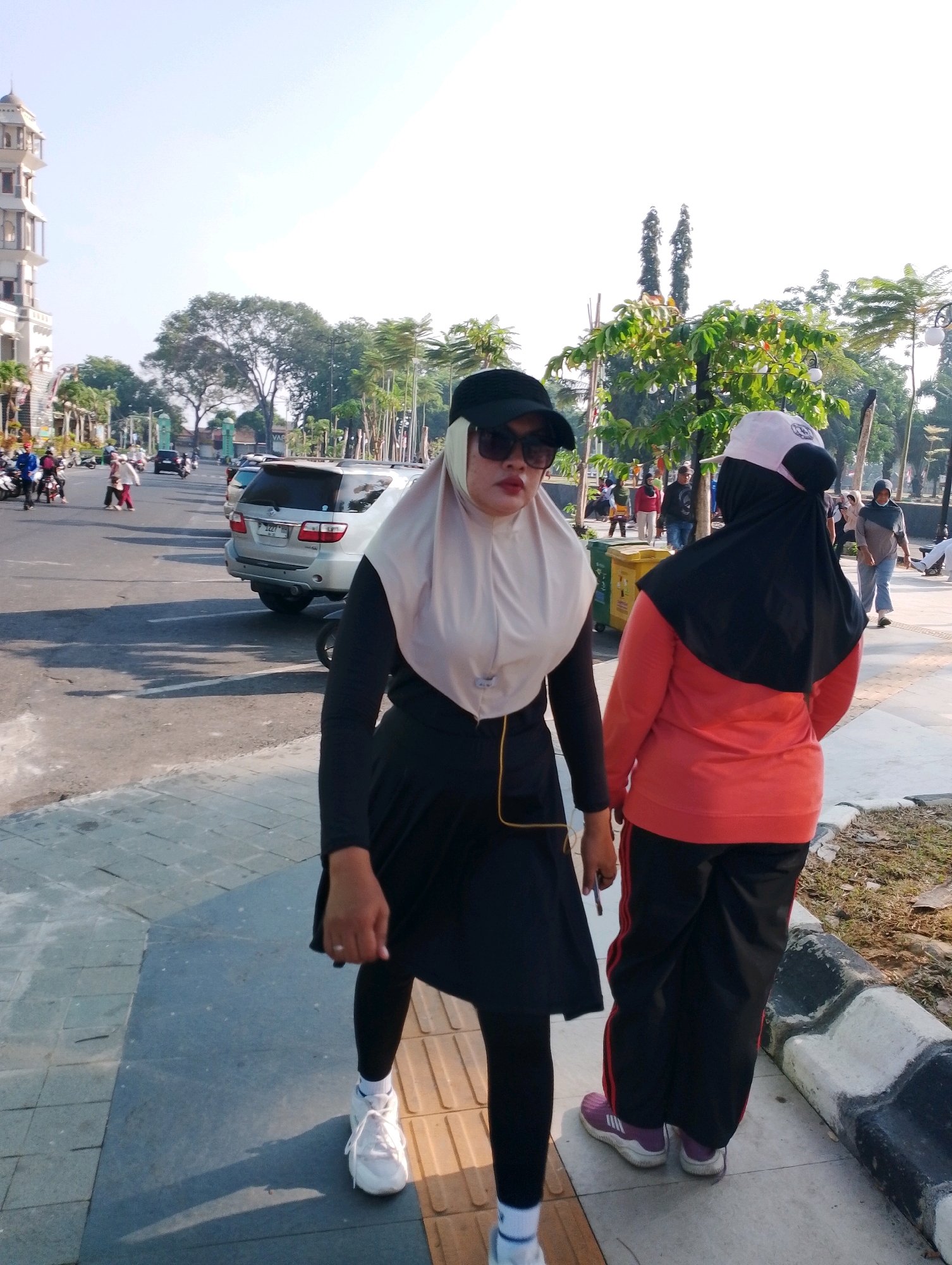 The image shows two women walking on a sidewalk next to a street in Probolinggo, Indonesia. The woman in the foreground is wearing a light-colored hijab, sunglasses, a black long-sleeved shirt, and a black skirt over black leggings. She has white sneakers on and is holding a pen in her right hand. The woman behind her is wearing a black hijab, a light pink cap, and a coral-colored long-sleeved shirt with black pants. She has purple sneakers on. The background shows a street with cars and motorcycles, trees, and a building with a tower on the left side. The sky is clear and blue. The scene appears to be a public area in the city, possibly a park or a street in a residential or commercial district.