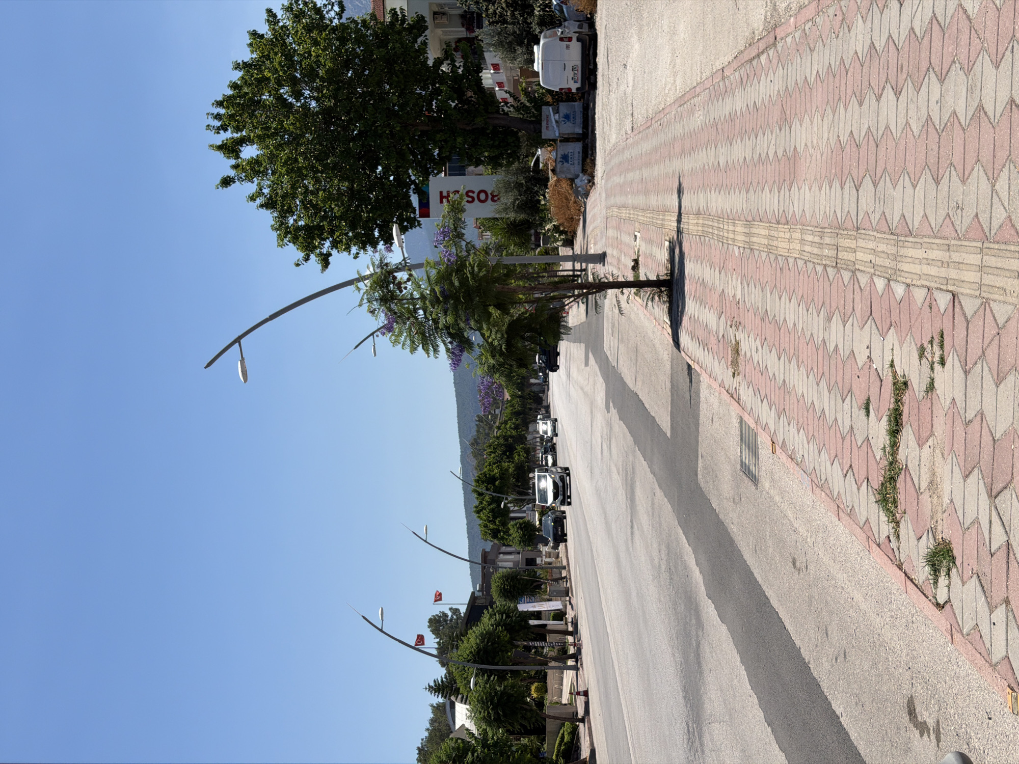 This media file depicts a street scene on a sunny day. The perspective is from the sidewalk, looking down a road lined with trees and streetlights. Several cars are parked or driving on the road, and a white van is visible in the background. The sidewalk is paved with a decorative pattern of pink and beige interlocking blocks. In the distance, there are buildings and hills under a clear blue sky. The overall mood is peaceful and ordinary, suggesting a typical day in a town or city. There is text visible on a large red and white sign which reads "BOSCH".