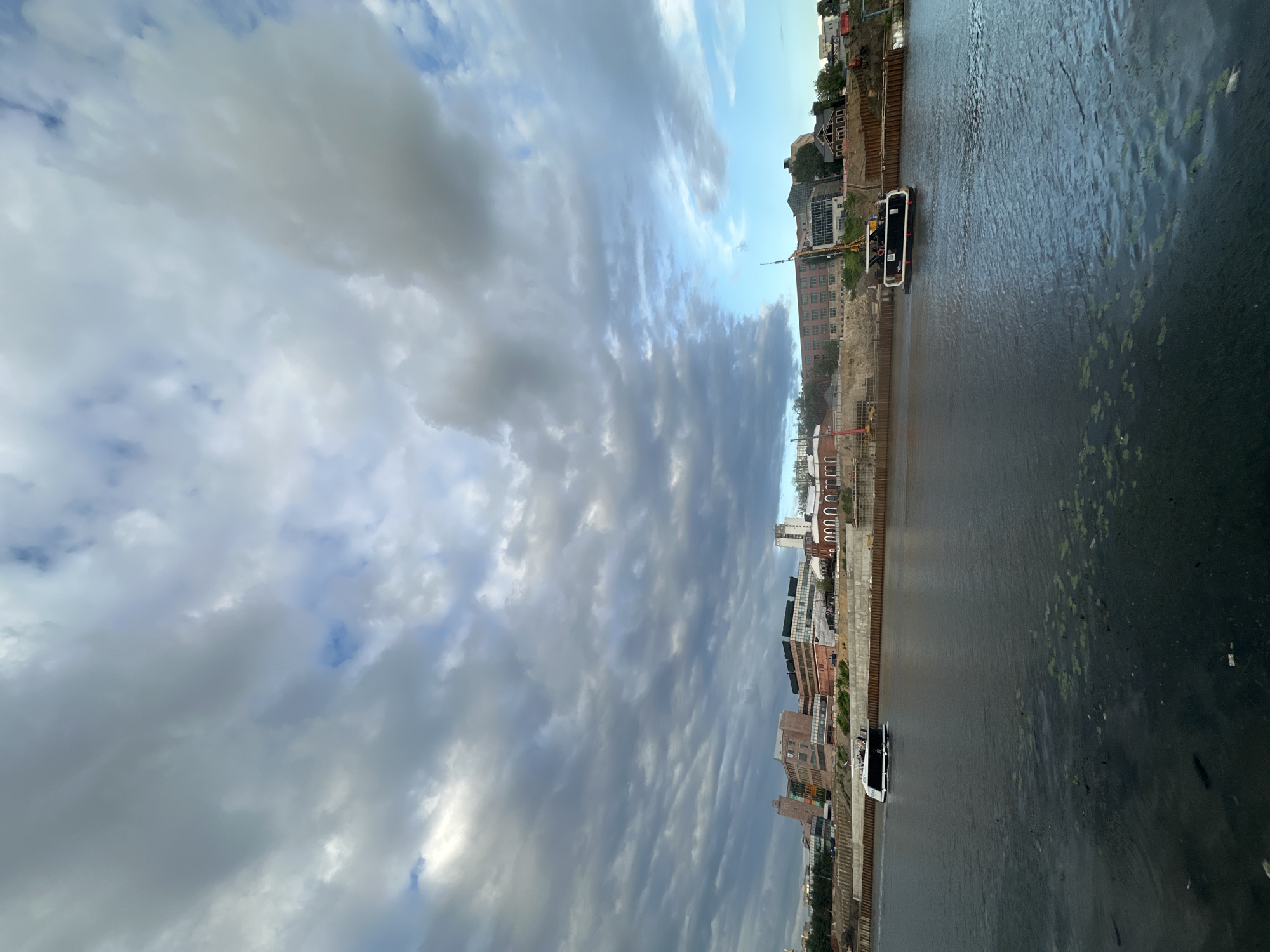 The image depicts a wide view of the Moskva River in Moscow, Russia, with a modern city skyline in the background. The sky is filled with dramatic, grey, and white clouds, hinting at impending weather. On the river, a sleek, white passenger boat is moving from left to right. The boat has several people on its upper deck. Along the riverbank, there are buildings of various architectural styles, some appearing to be undergoing construction or renovation, indicated by cranes and scaffolding. The water of the river is dark and somewhat murky, with patches of green algae visible on the surface. The overall scene suggests a late afternoon or early evening on a cloudy day in an urban environment.