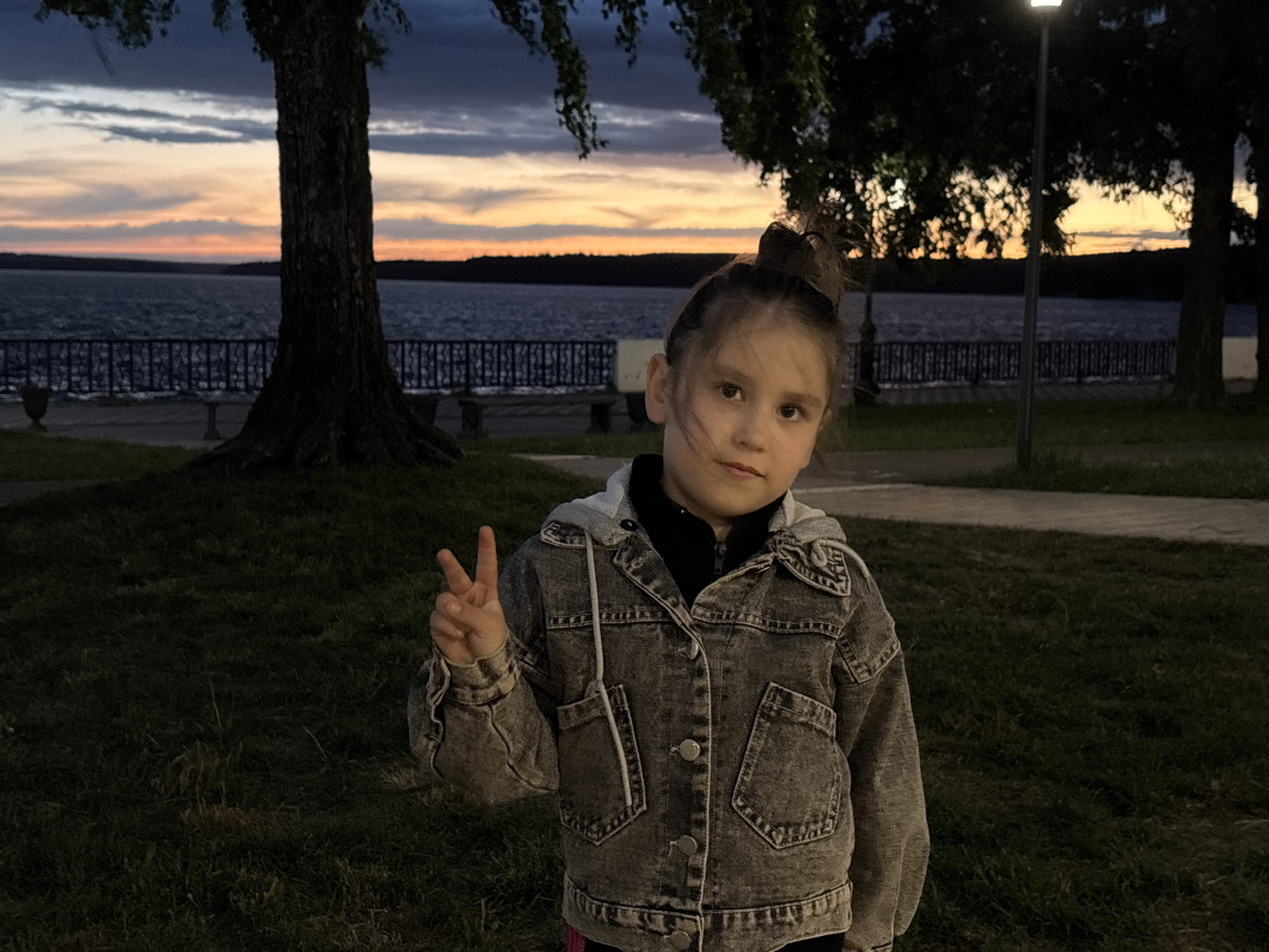 A young girl is standing on a grassy area next to a paved walkway with a body of water, a black wrought iron fence, and trees in the background. The girl is wearing a denim jacket with a gray hoodie underneath, and she is making a "peace" sign with her left hand. The sky in the background shows colors of orange and purple, indicating either sunrise or sunset. In the background, the location is identified as the Votkinsk Reservoir, on the embankment of the city of Votkinsk, Russia, close to the pier area and the Peter Tchaikovsky monument.