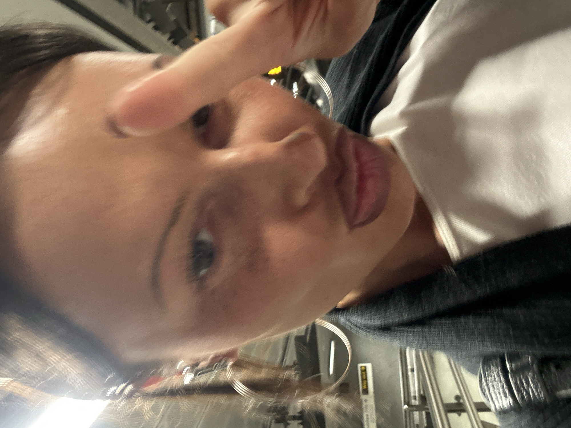 A close-up selfie captures a person, likely female, from a low, upward-looking angle, with the image rotated 90 degrees counter-clockwise. The subject has dark hair, visible on the left, and a warm skin tone. They are looking towards the camera with a playful expression, featuring slightly pursed lips with dark lipstick and one eye visible. A finger from their right hand is raised, pointing or gesturing towards the camera, partially obscuring their forehead.
The person is wearing a dark, possibly textured or denim-like outer garment over a plain white top. Large, thin metallic hoop earrings are visible near their ear on the right side of the frame, catching reflections.
The background suggests an indoor public transport environment, likely a subway or metro station, consistent with the London, United Kingdom context. Visible elements include metallic bars or railings, and reflections from overhead artificial lighting. A yellow and black sign in the background, to the right of the subject's head, displays the text "Way out", further confirming a station setting. The overall lighting is artificial and somewhat soft, illuminating the subject's face while creating highlights on the metal elements. The activity is a casual selfie, conveying a candid moment.