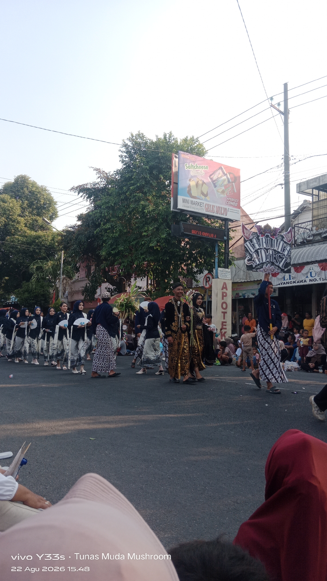 A street procession is taking place in Genengan, Indonesia. A group of people dressed in traditional Javanese attire, including batik skirts and patterned head coverings, are walking in formation. Some are holding fans. A couple in ornate gold and black clothing, also in traditional Javanese style, are walking at the front of the procession, appearing to lead it. The procession is on a paved street with onlookers seated on the side. In the background, there are trees, buildings, a billboard advertising "Saltcheese Condo," and streetlights. The sky is clear and bright. The timestamp "22 Agu 2026 15.48" and "vivo Y33s. Tunas Muda Mushroom" are overlaid on the image.