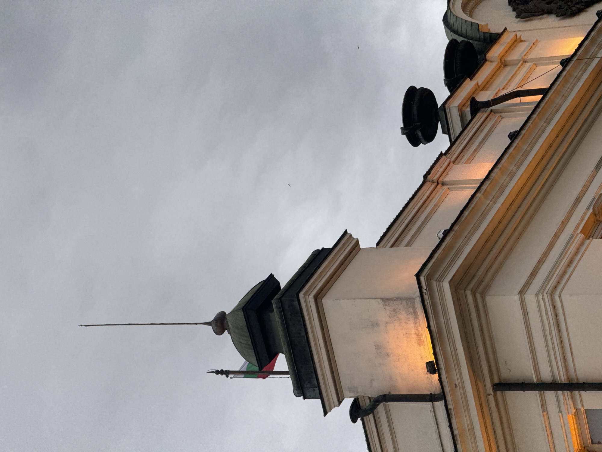 The image shows a low-angle view of architectural details from a building in Sofia, Bulgaria, against a gray, overcast sky. The building is characterized by its white facade with dark trim. The structure appears to have classical elements with vertical pilasters and layered cornices. A green and red flag hangs from a turret-like top of the building. A spire protrudes upward into the sky from the turret. Circular dark structures are mounted on the side of the building. The overall scene suggests an urban setting with historical buildings in Sofia.