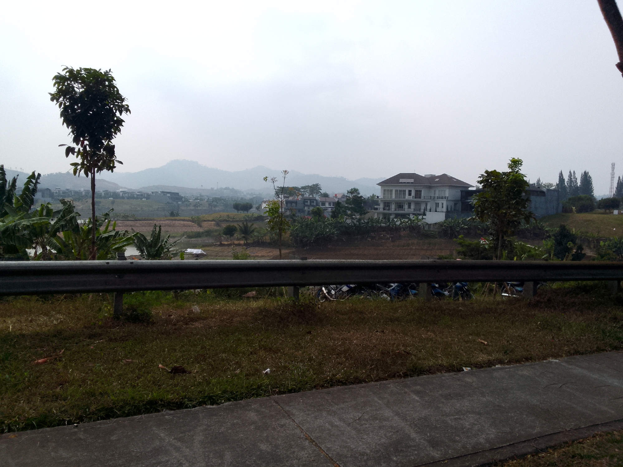 The image depicts a landscape in Bojonghaleuang, Indonesia, featuring a residential area with modern architecture, rolling hills, and a cloudy sky. In the foreground, a metal guardrail runs horizontally across the frame, separating a paved walkway from a grassy embankment. Several motorcycles are parked beyond the guardrail on the grass. A tall, slender tree with dark green foliage stands to the left, with banana plants visible behind it. In the middle ground, a large, multi-story white house with numerous windows and a prominent balcony is situated amidst other buildings and trees. The background shows a hazy view of distant mountains and more residential development. The overall impression is of a suburban or semi-rural environment under overcast conditions.