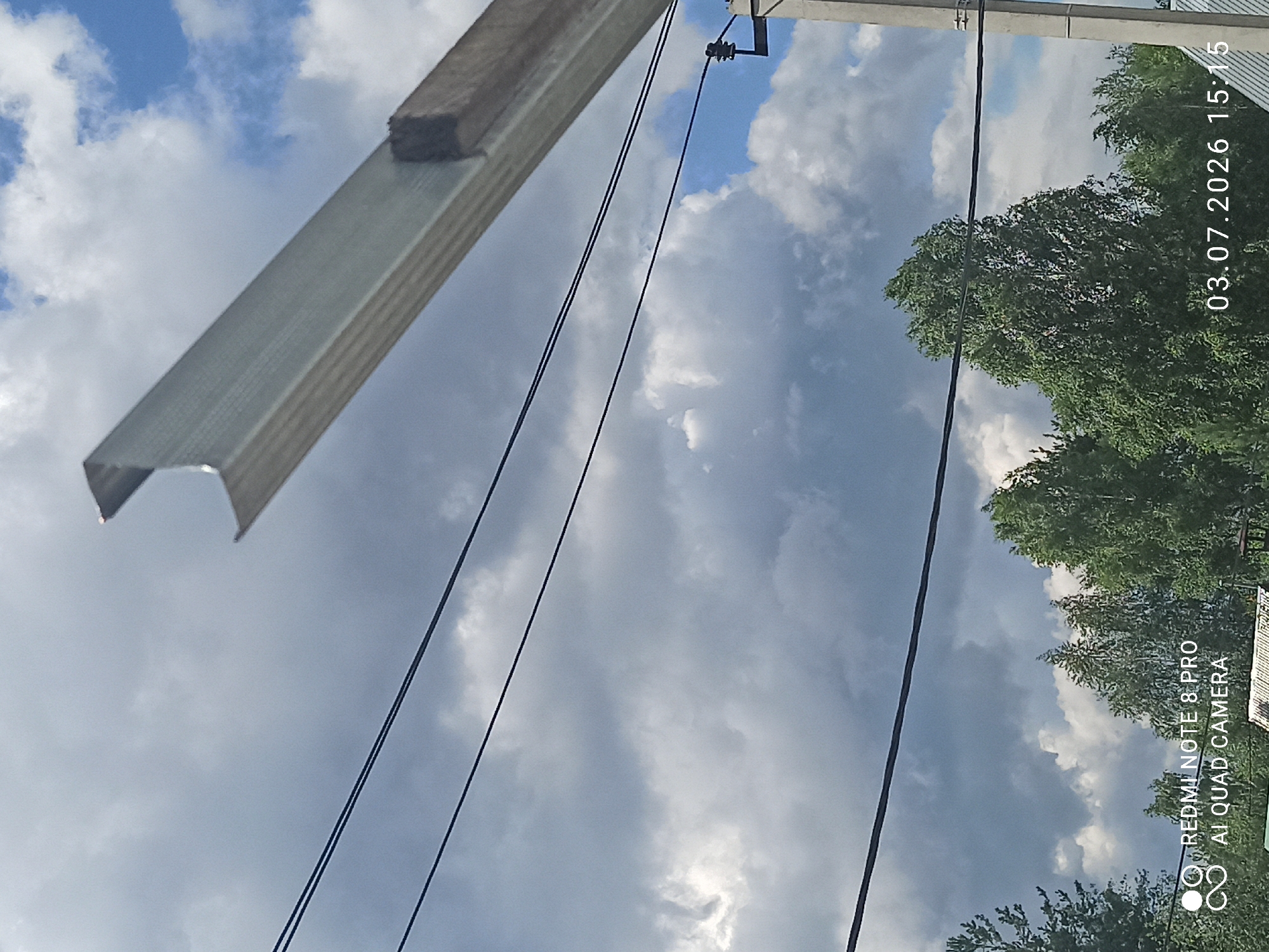 The image depicts an outdoor scene with a cloudy sky overhead. In the foreground, a metallic beam is angled from the upper right to the lower left, supported by a wooden block. Several dark power lines are strung horizontally across the frame. Below the power lines, lush green trees are visible, with some appearing taller and fuller than others. In the lower left corner, text overlays indicate "REDMI NOTE 8 PRO AI QUAD CAMERA", and in the lower right corner, a date and time stamp reads "03.07.2026 15:15". A concrete pole with electrical insulators is present on the right side of the image. The overall scene appears to be a somewhat overcast day in a semi-rural or suburban environment, possibly Sovetskiy, Russia, given the location tag. There are no people or specific actions depicted.