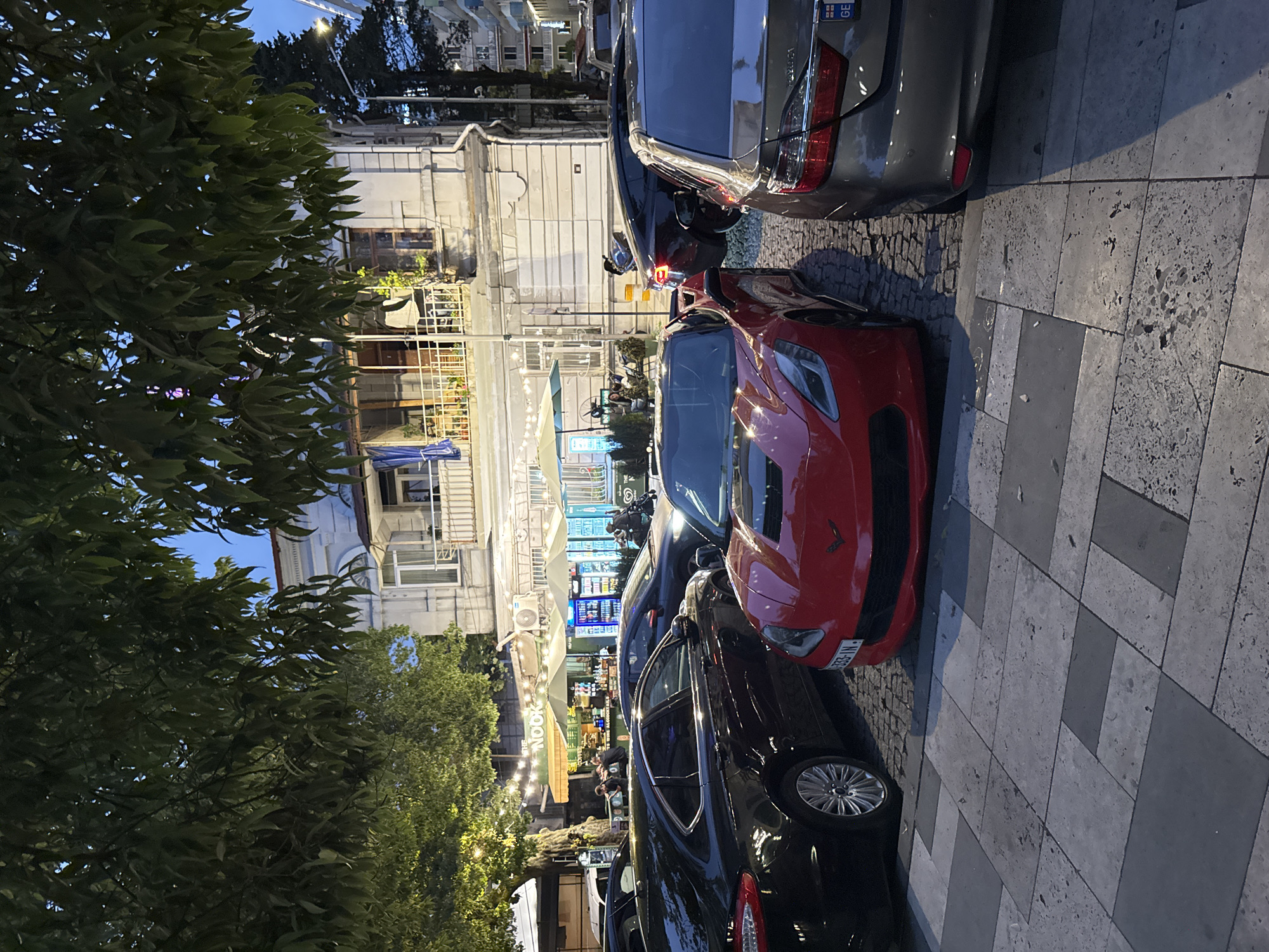 This image depicts a street scene in Batumi, Georgia, likely in the evening or at dusk, as indicated by the low light and artificial illumination from storefronts and string lights. The foreground is dominated by several parked cars, including a striking red sports car and a black sedan. Beyond the cars, a building with multiple levels and balconies is visible, with various storefronts and seating areas in front of it, suggesting a commercial or dining establishment. Some trees with lush green foliage are present, framing the scene. String lights are strung across some of the outdoor seating areas, contributing to the evening ambiance. People are present, though mostly indistinct, appearing to be patrons or staff at the establishments. The ground is paved with large, irregular stones. Visible text includes "NOOK" on a green awning and license plates on the cars, one of which appears to be "GE" followed by numbers and letters, consistent with Georgian license plates. The overall atmosphere suggests a lively, albeit quiet, evening in a developed urban area.