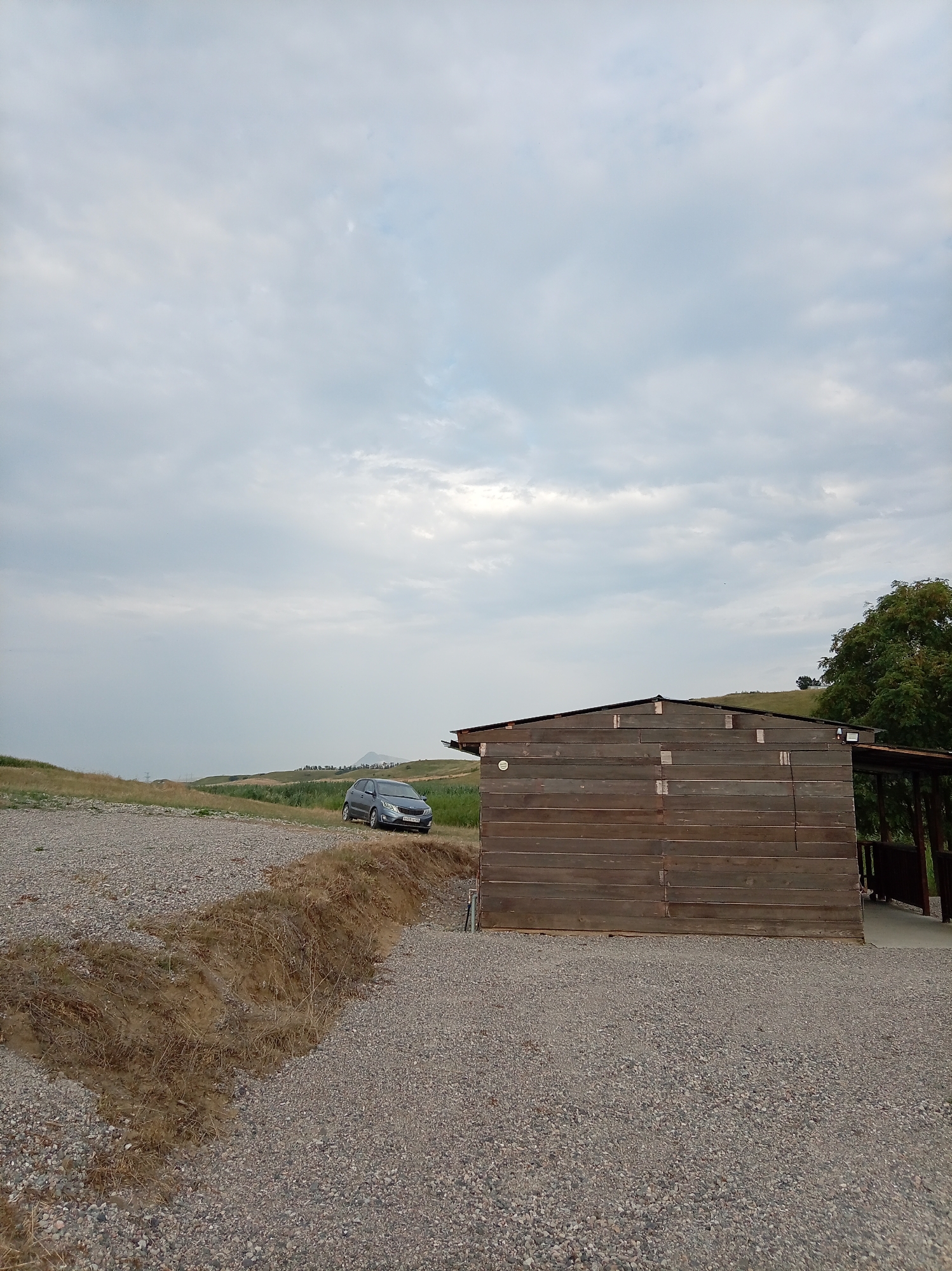 The image depicts an outdoor scene near a wooden structure. In the background, rolling hills are visible under a cloudy sky. A grey car is parked on a gravel surface to the left of the wooden building. The wooden structure is of modest size and appears to be a shed or a small outbuilding, with horizontal wooden planks forming its walls. A gravel pathway leads away from the structure and towards the car. There is dry grass and soil in a trench-like area in the foreground. The scene suggests a rural or semi-rural setting, potentially on the outskirts of Гражданское, Russia.