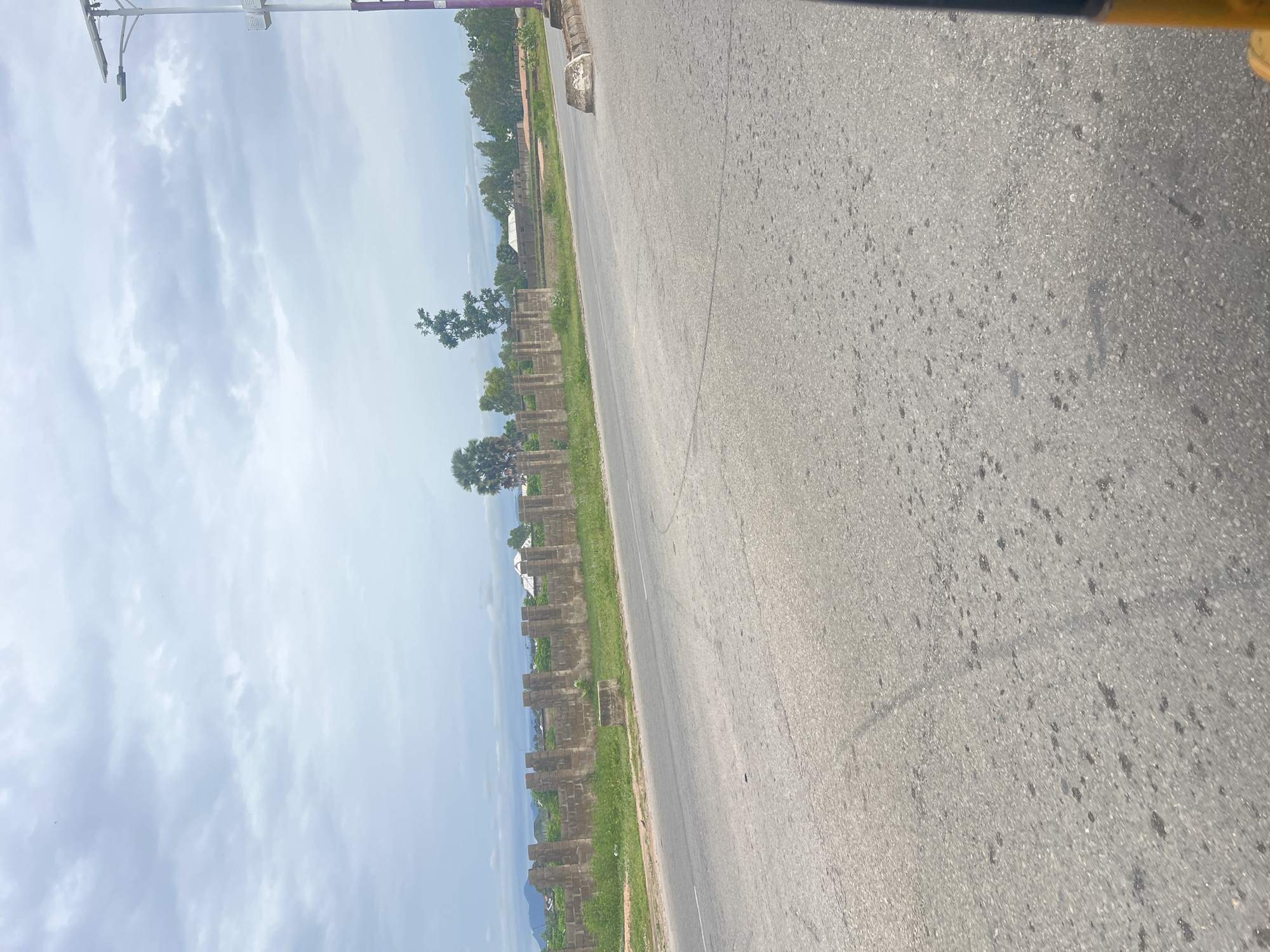 This media file depicts a roadside scene in Jalingo, Nigeria. The image is oriented vertically, with the road occupying the right side of the frame and the sky and distant landscape on the left. The road appears to be paved with asphalt and shows signs of wear, with a white line marking the lane. To the left of the road, there is a grassy verge followed by a row of stone pillars, suggesting the remnants of a fence or boundary. Beyond the pillars, there are trees and some buildings, indicating a semi-rural or developing area. In the far distance, a body of water, likely a lake or river, is visible, with hills or mountains in the background. The sky is partly cloudy, with soft, diffused light, suggesting it could be morning or late afternoon. There are no people or vehicles visible in the image. The overall impression is one of a quiet, possibly undeveloped, area near a body of water.