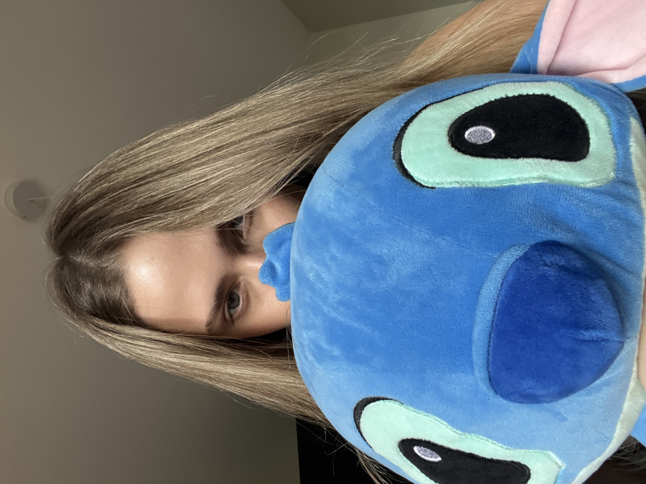 An indoor, head-and-shoulders portrait features a young adult female with long, blonde hair parted down the middle. She is holding a large blue plush toy, identified as the character Stitch, in front of her face, partially obscuring her features. Her blue eyes are visible peering over the top of the plush toy, looking directly at the viewer. The plush toy's prominent embroidered eyes, with black and light blue coloration, and its dark blue nose are clearly visible. A small pink section, likely an ear of the toy, is visible on the far right. The background consists of a plain, light-colored ceiling with a single, circular white light fixture mounted on it in the upper right portion of the frame. There are no discernible visual elements in the image to indicate a specific location within Saint Petersburg, Russia.