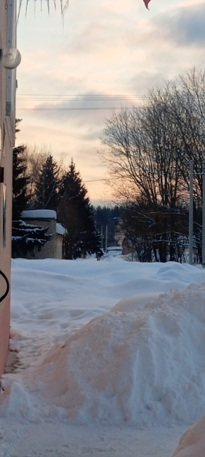 The image portrays a winter street scene in Красный Маяк, Russia, under an overcast sky exhibiting soft pale orange, pink, and blue tones. The foreground features a substantial amount of undisturbed white snow, forming deep drifts. On the far left, a segment of a pale-colored building is visible, equipped with a spherical metallic light fixture. Multiple clear icicles hang from an unseen structure above the building on the left. A cleared or less snow-covered path runs parallel to the building on the immediate left.
The midground reveals a snow-covered thoroughfare or path receding into the distance, bordered by utility poles carrying overhead wires. To the left, a small, light-colored building with snow accumulating on its roof is partially visible. The scene is populated by bare deciduous trees and dark coniferous trees, particularly evident in the midground and background, where they form denser clusters. A small, reddish, triangular object is barely discernible against the sky in the upper right corner. No individuals, animals, or active subjects are present in the scene.