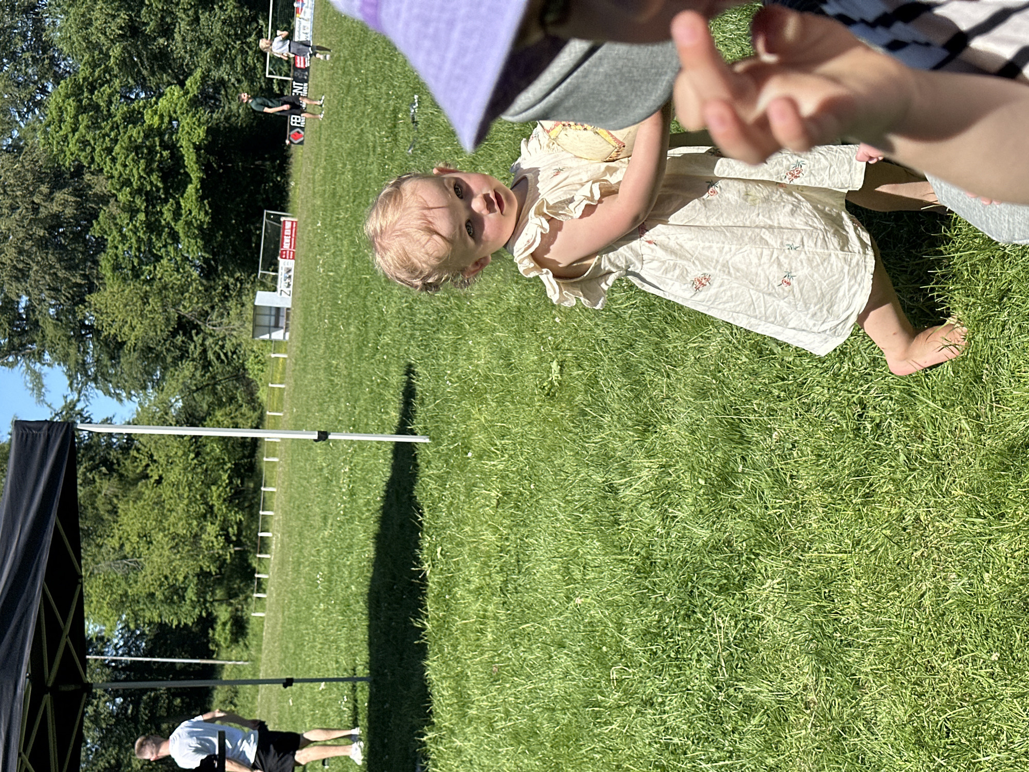 In Grünberg, Germany, a young, fair-haired child wearing a light-colored dress stands barefoot on a grassy field. An adult wearing a striped shirt and a hat is interacting with the child. In the background, there is a line of people, a soccer goal, and trees. A black tent is visible in the foreground, and a man is standing nearby.