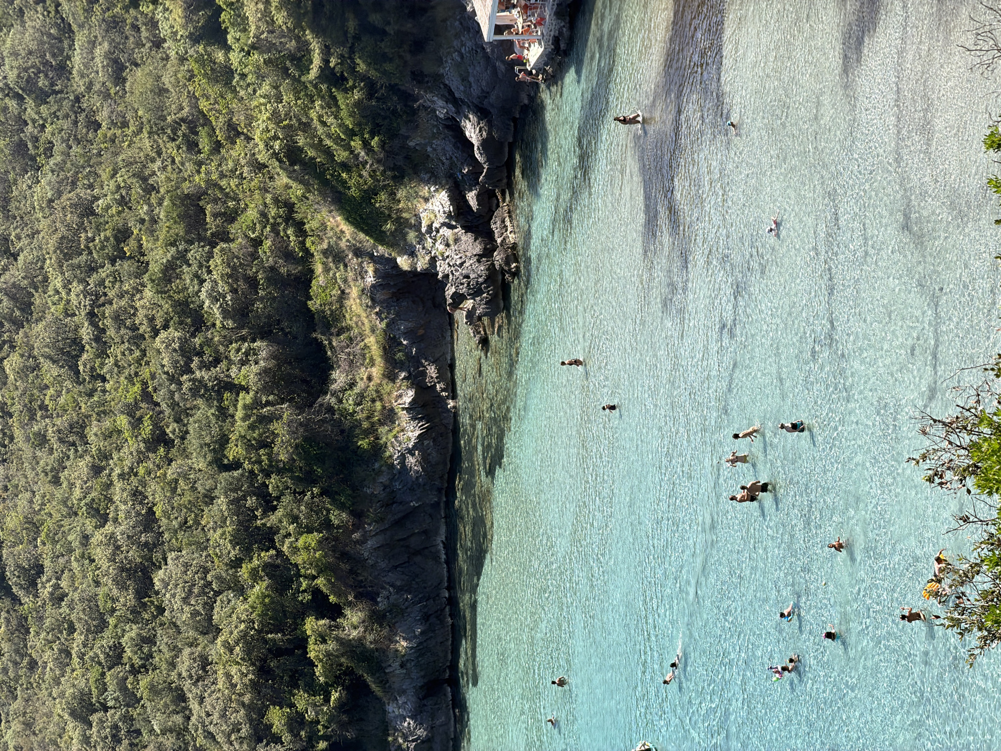 This is a high-angle, medium shot of a bay at Dubraljevina, Montenegro, on a sunny day. The water is a clear, vibrant turquoise, revealing the sandy bottom in shallower areas. Numerous people are enjoying the water. Some are swimming, others are wading in the shallows, and a few are standing on rocks near the shore. In the background, a lush, green hillside covered in dense vegetation slopes down to the rocky coastline. On the right side of the bay, a small, white structure with an orange roof, possibly a beach bar or restaurant, is visible with people seated at tables. The scene depicts a peaceful and idyllic summer day at the coast.