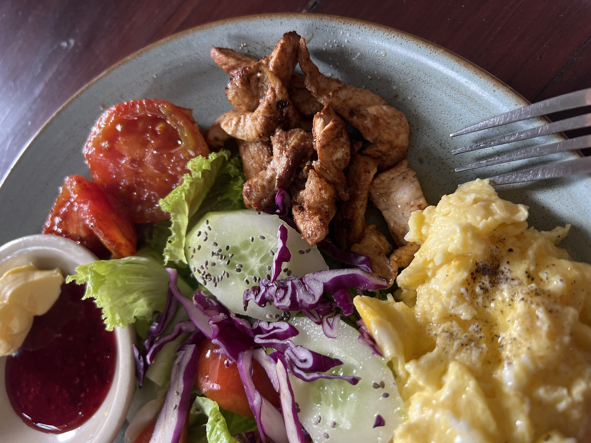 A close-up shot shows a plate of food, likely taken in North Kuta, Indonesia, given the food presentation. The plate contains scrambled eggs sprinkled with black pepper, stir-fried chicken pieces, slices of tomato, cucumber (with what appears to be chia seeds), red cabbage, and lettuce. A small dish on the side holds two sauces, one yellow and one red. A portion of a metal fork is visible on the right side of the plate. The plate itself is a muted blue-green color with a brown rim. The plate is set on a dark brown surface.