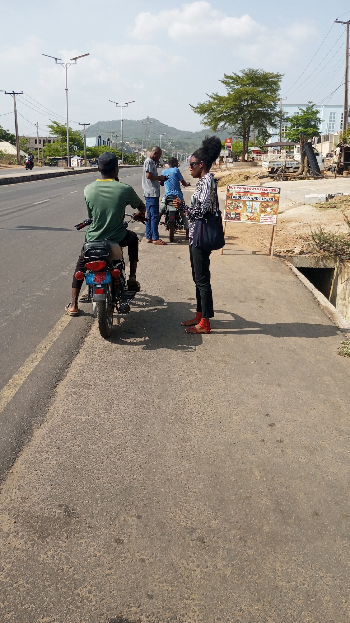 The scene is in Adankolo, Nigeria. A man in a green shirt sits on a motorcycle parked on the sidewalk next to a road. A woman in a striped shirt and black pants stands facing him, seemingly engaged in a conversation. Two other men are visible near another motorcycle further down the sidewalk. A sign that says "Buy Your Fowls Feed Here" is partially visible. A road is visible on the left side of the image. In the background, there is a mountain and some buildings. Overhead power lines and street lights are present.