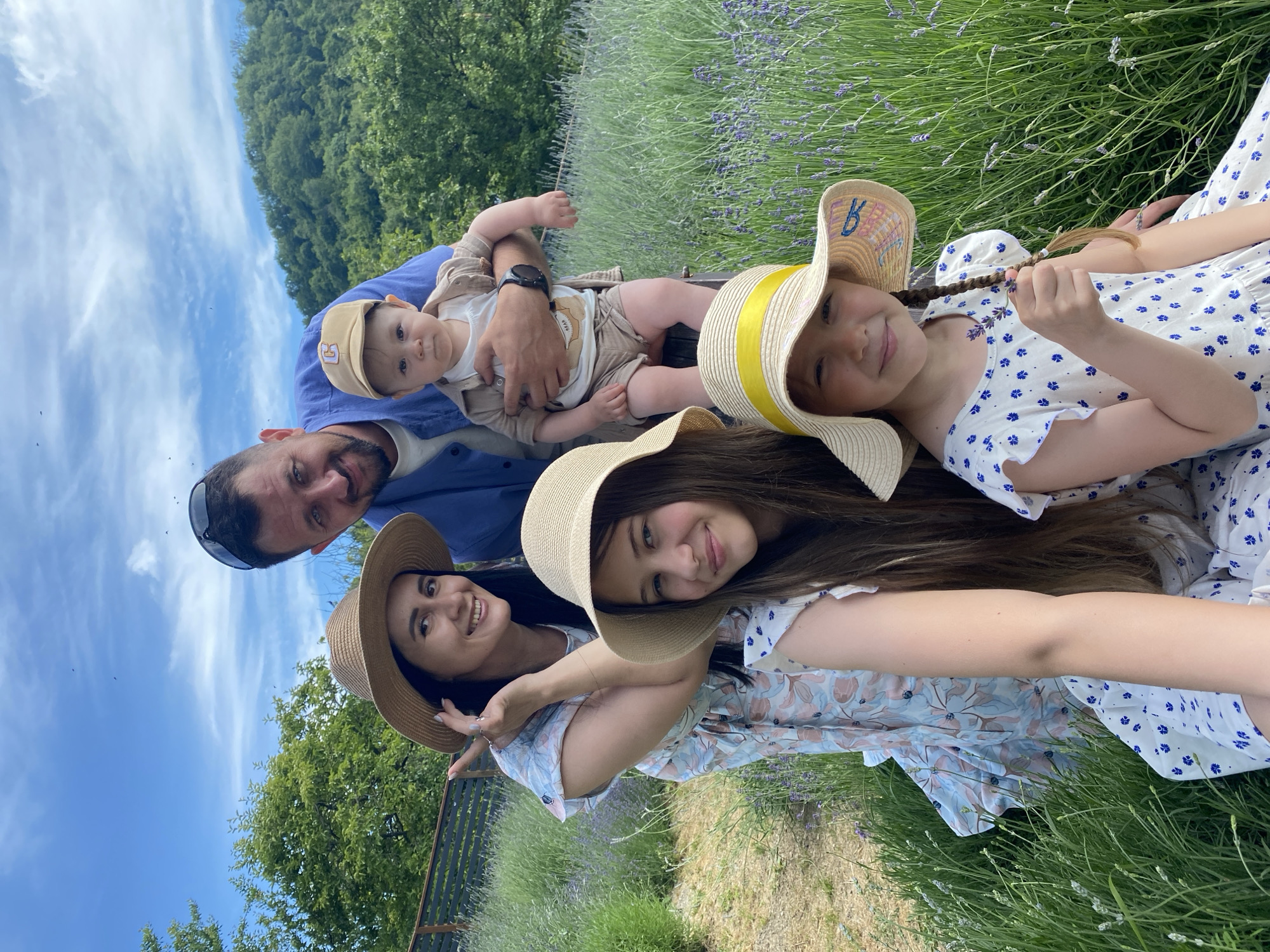 In Verkhneye Uchdere, Russia, on a bright, sunny day, a family of five poses for a selfie in a lavender field. The family consists of two adults and three children. The father, wearing a blue shirt, holds a baby in his arms. The mother, in a floral dress and a straw hat, stands to the left. Two daughters, one in the foreground wearing a white dress with blue flowers and a straw hat with a yellow ribbon, and another behind her, also in a straw hat, complete the group. The background features lush green trees and a blue sky with scattered clouds. The ground is covered in tall green grass and purple lavender flowers. The overall scene depicts a happy family outing in a natural setting.