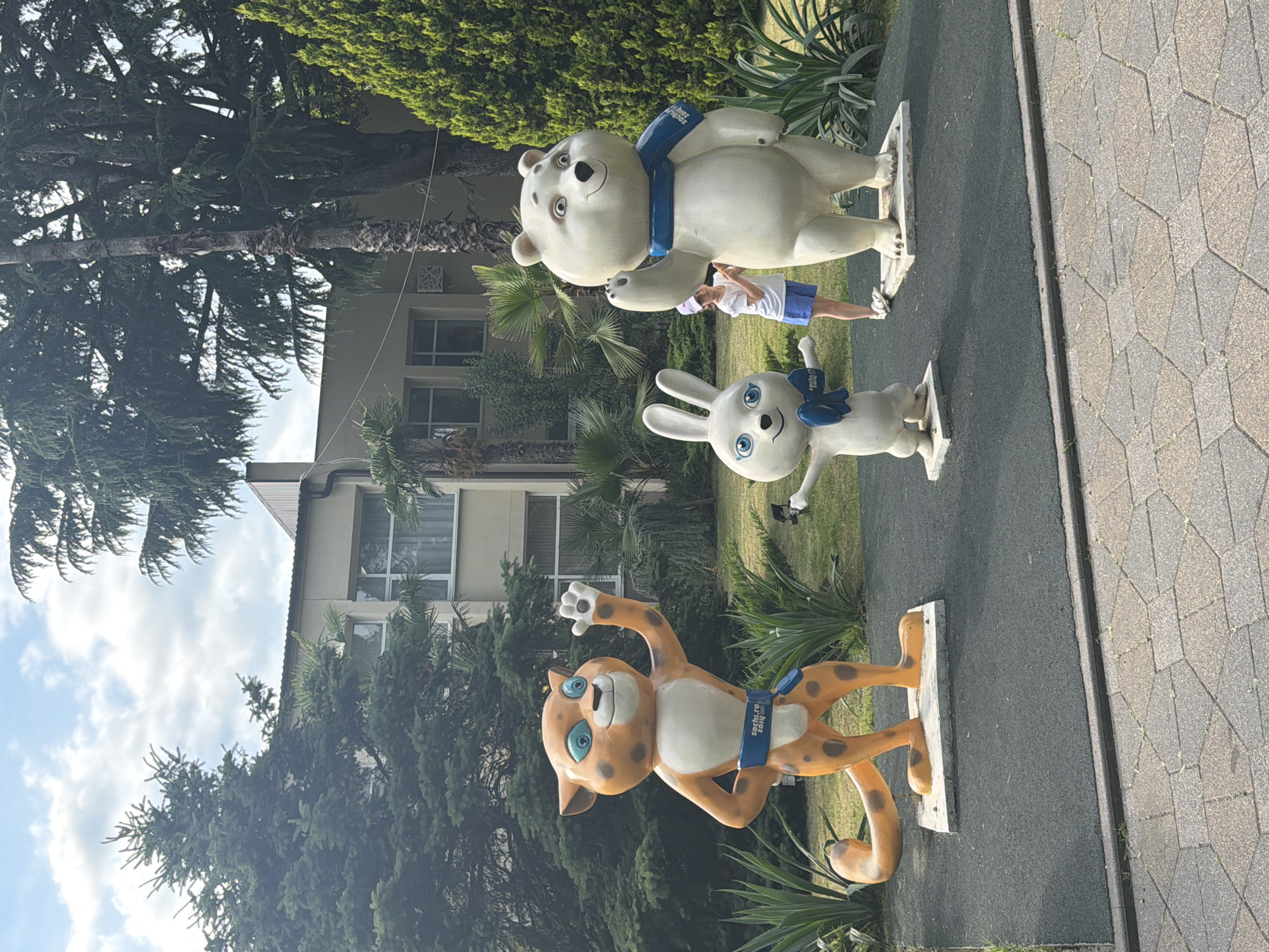 The image displays three statues of Olympic mascots in an outdoor setting. From bottom to top, the statues depict a leopard, a rabbit, and a polar bear. The leopard, dressed in an orange and white spotted costume with a blue band, waves its paw. The rabbit is white with blue accents. The polar bear is white and also has a blue scarf. A young person wearing a white shirt and blue shorts stands between the polar bear and the rabbit, appearing to interact with the statues.
The statues are placed on a dark grey surface, possibly a pathway or park area, with a paved walkway to the right. Lush green trees and foliage surround the scene. In the background, a building with multiple windows is visible, suggesting an urban or resort environment. The sky is partly cloudy, indicating fair weather. The time of day appears to be daytime, with bright lighting. The blue bands on the statues are inscribed with "Sochi 2014". The overall impression is one of a sunny day at a location commemorating the Sochi 2014 Winter Olympics.