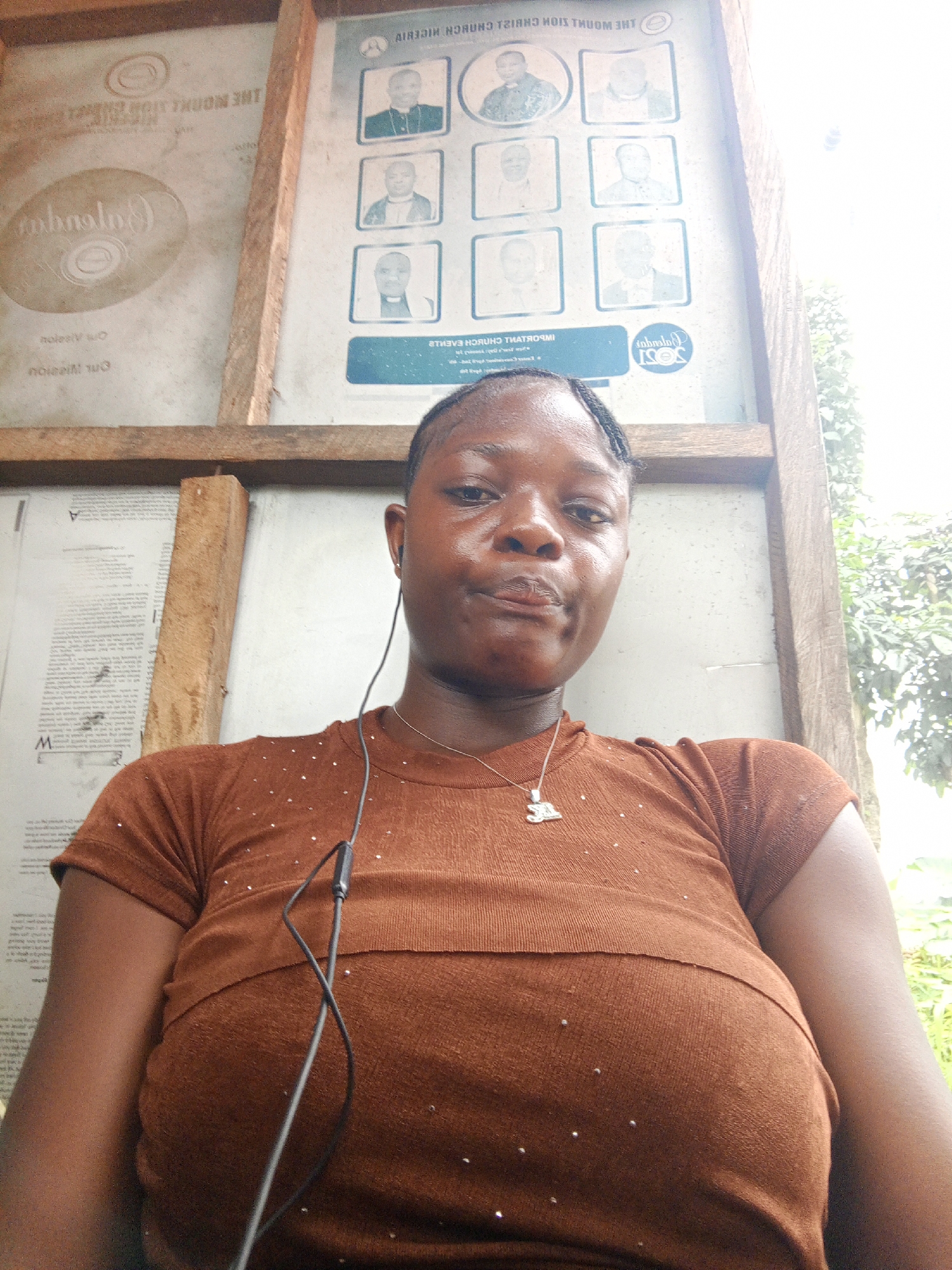 The image shows a woman in a brown shirt standing in front of a bulletin board. The bulletin board contains various postings and announcements, including images of religious leaders. The board also displays the name "The Mount Zion Christ Church Nigeria." The setting appears to be outdoors, with trees visible in the background. The location is Mbribit, Nigeria. The woman has a necklace with a pendant and a wired earphone in her ear.
