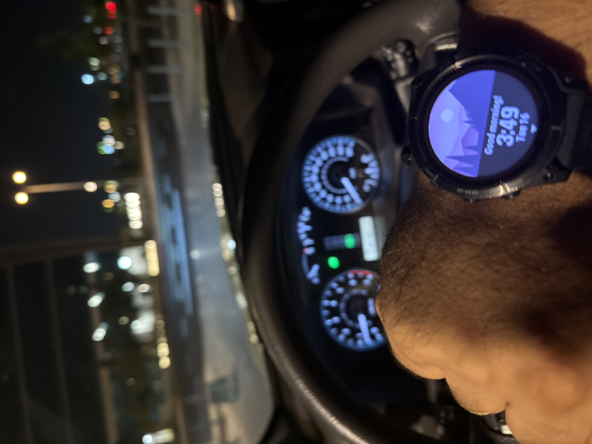A first-person perspective shot shows a hand wearing a smartwatch and resting on a car's steering wheel. The smartwatch displays "Good morning! 3:49 Tue 16" over a minimalist mountain landscape. The car's dashboard, with illuminated gauges and a digital display, is visible in the background, as is a blurry night-time cityscape with streetlights and building lights. The environment appears to be urban, possibly a parking lot or roadside at night.