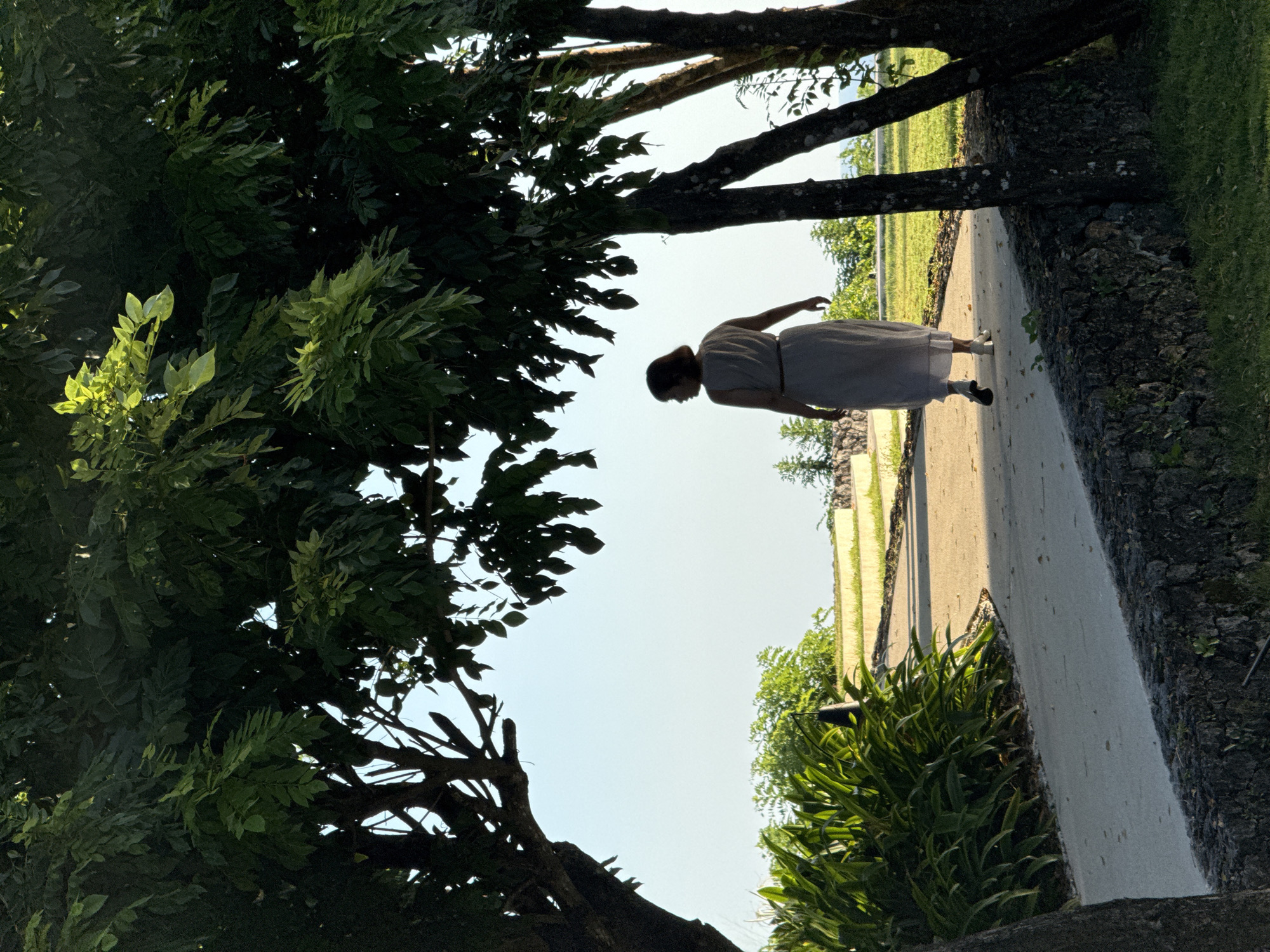 A woman in a light-colored dress is walking along a paved path in a garden in Kuta Selatan, Indonesia. The path is lined on one side by a low stone wall, and on the other side by a manicured garden with green grass and hedges. The woman is walking away from the camera, and her silhouette is visible against the bright sky. Two trees are in the foreground. The tree on the left has dense green leaves that block some of the sun, but the tree on the right is further back and has less dense coverage.