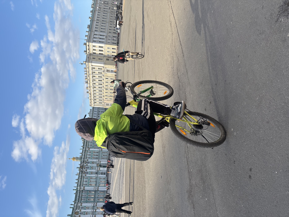 The image displays Palace Square in Saint Petersburg, Russia, under a partially cloudy blue sky. In the foreground, a person, viewed from behind, is performing a wheelie on a bright green and yellow bicycle. This individual wears a neon yellow-green jacket, a dark hooded garment with a backward cap, dark pants with white side stripes, and dark sneakers, carrying a black backpack with orange zipper details. The bicycle features disc brakes and orange spoke reflectors on the rear wheel. To the mid-ground right, another individual rides a dark-colored bicycle, wearing a dark hoodie with a red graphic. The background features the turquoise and white Baroque-style Winter Palace (Hermitage Museum) on the left, distinguished by its columns, ornate facade, and a prominent golden spire. To the right, part of the light-colored General Staff Building is visible. Numerous pedestrians are present across the vast paved square, with vehicles identifiable in the far distance on the right.