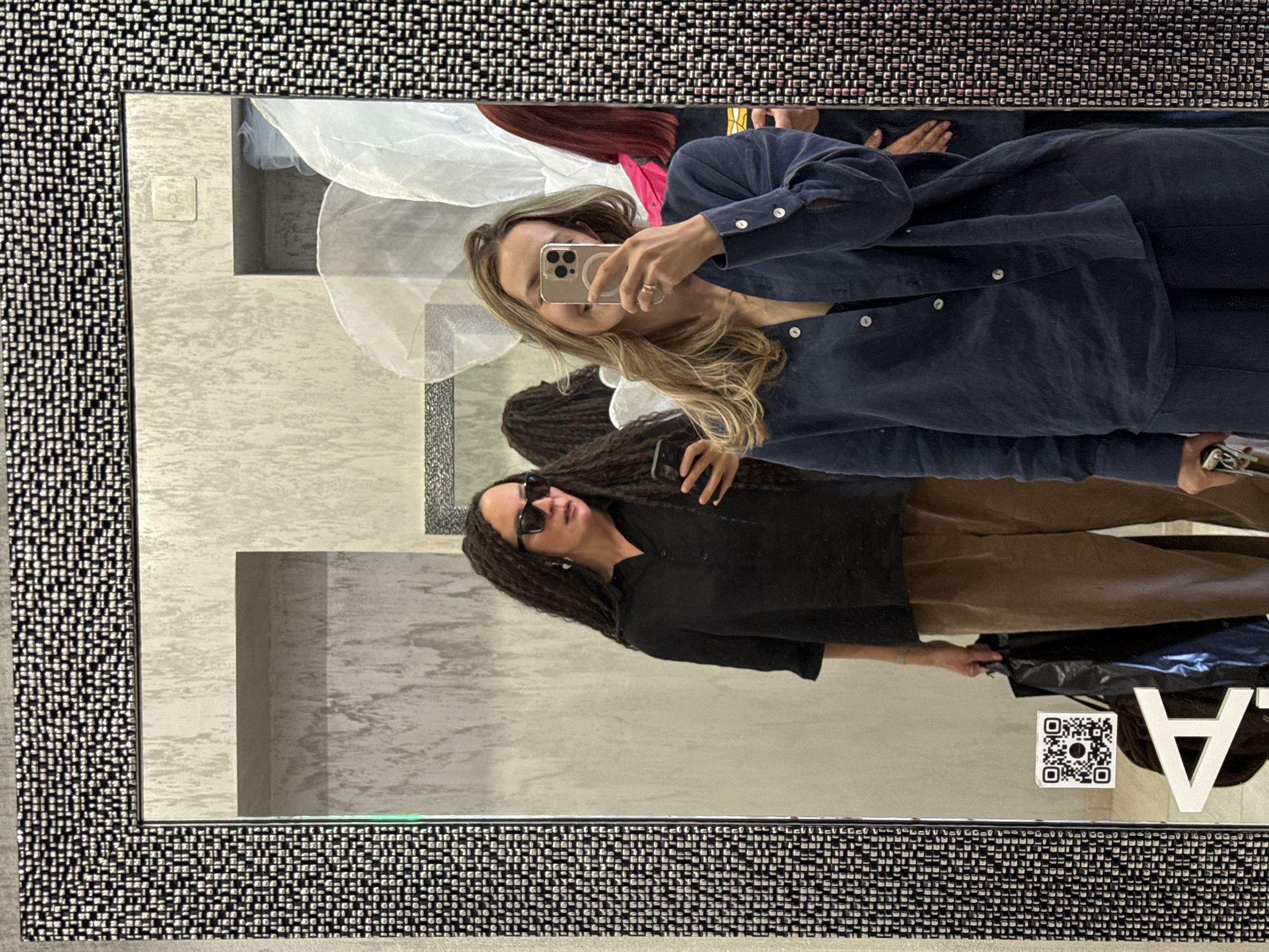 A mirror reflects two women taking a photo, likely in a retail environment. The first woman, situated at the top of the frame, is holding a smartphone and appears to be taking the photo. She has long blonde hair and is wearing a navy blue button-down shirt. The second woman, positioned below, has long dark hair, sunglasses, and a black top. She is holding a black shopping bag. The background shows interior elements, including sections of wall. A QR code and a white "A" are visible near the bottom edge of the mirror. The location could be a retail store or shop. No specific evidence indicates this is Chaikovsky, Russia.