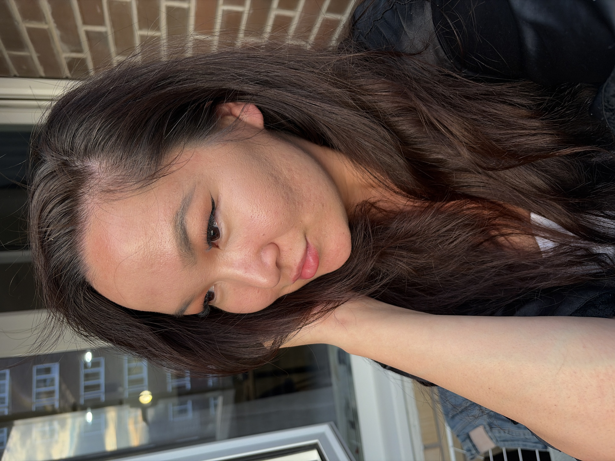 The image shows a close-up, side profile of a woman with long, dark brown hair. She is looking to her left with her hand near her face. The background suggests an urban setting with a brick wall on the right and a building with multiple windows reflected in a large glass pane on the left. The lighting indicates it is either early morning or late afternoon. The woman is wearing a black top, and a glimpse of denim is visible on her left side. The image does not contain any information that would allow for identification of the location as Izhevsk, Russia.