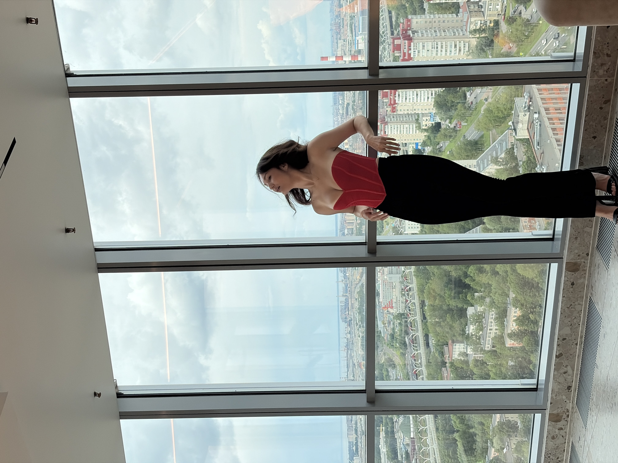 A woman stands on a balcony, leaning against a large window that overlooks a cityscape. She is wearing a red corset and a long black skirt, with her head tilted back and eyes closed. The window frames a panoramic view of Saint Petersburg, Russia, with its characteristic red-roofed buildings, lush green trees, and a glimpse of the Gulf of Finland in the distance. The sky is overcast with dramatic clouds, suggesting a possibly stormy or dynamic weather condition. The interior of the room is modern, with a sleek ceiling and patterned flooring. There are two visible fire sprinklers on the ceiling, indicating a commercial or residential building equipped with safety systems. The perspective from the high vantage point emphasizes the vastness of the urban landscape and the sheer scale of the city.